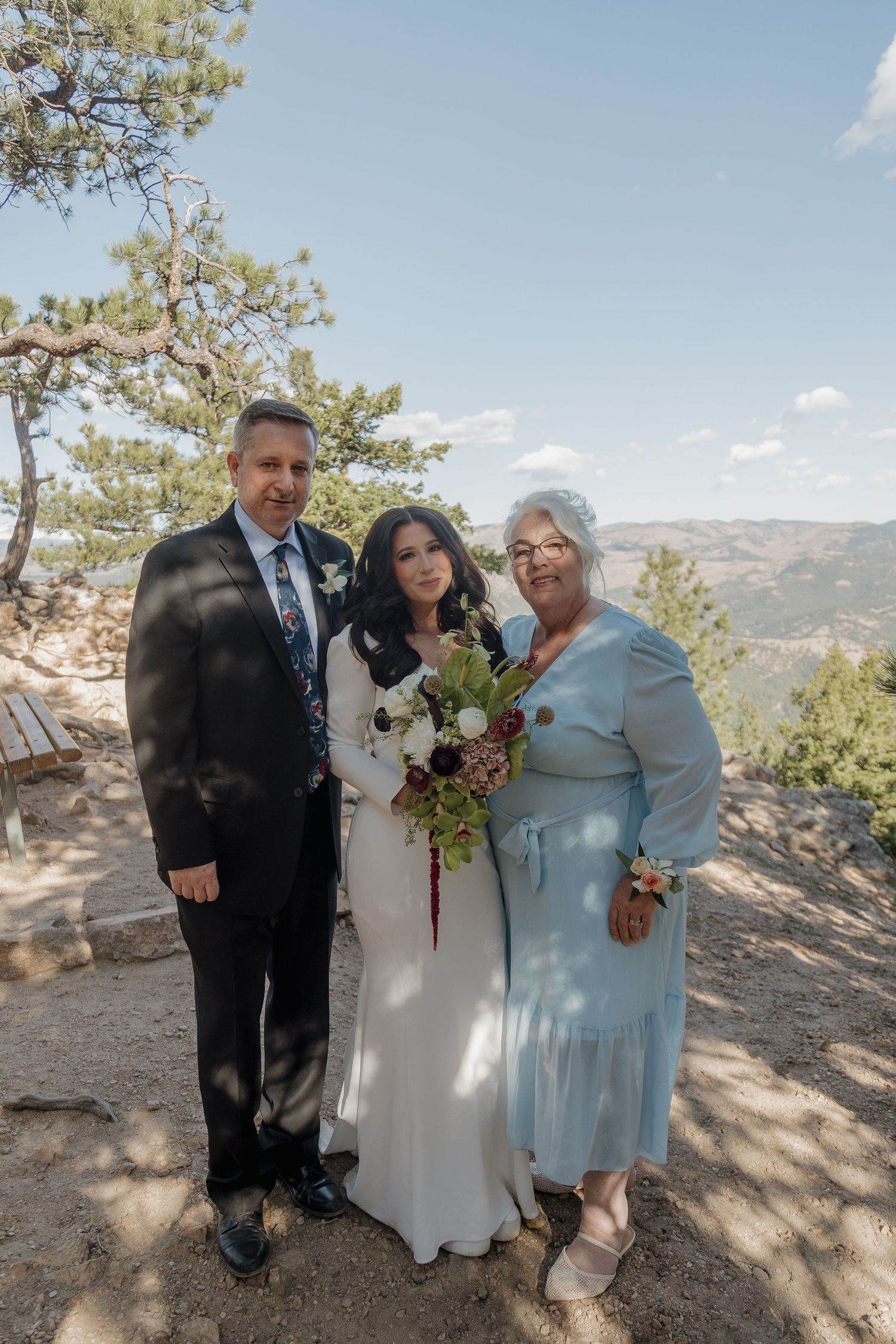 Bride with bouquet poses with two family members on mountain overlook under blue sky.