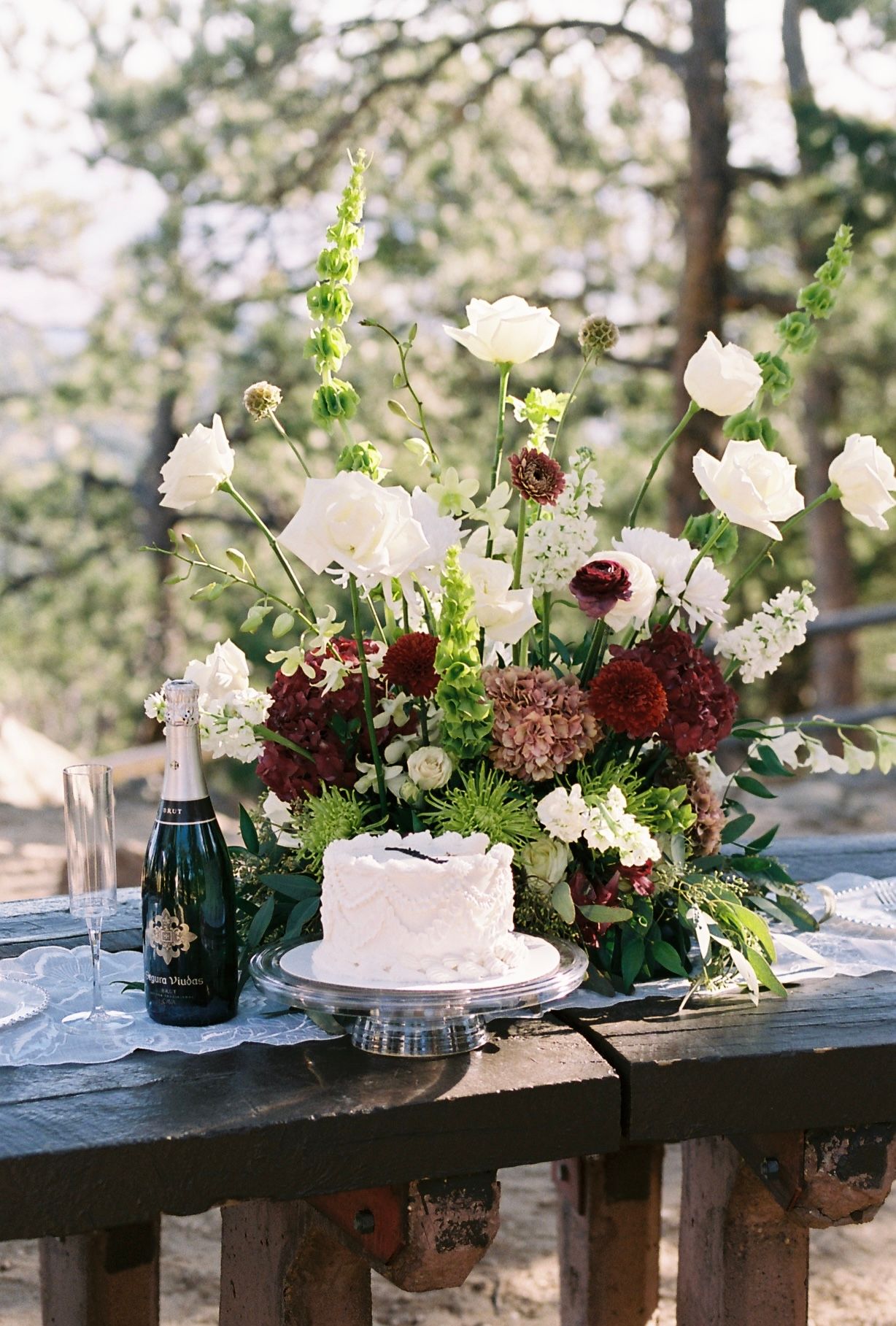 Elegant wedding cake with white floral arrangement and champagne bottle on rustic outdoor table.