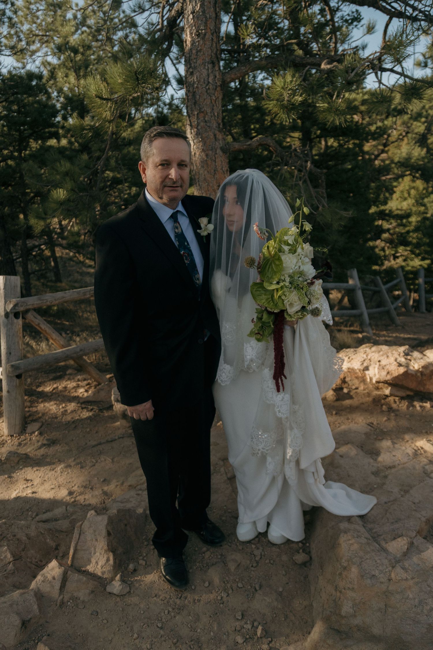 Bride in white gown and veil poses with suited man among pine trees at outdoor wedding.