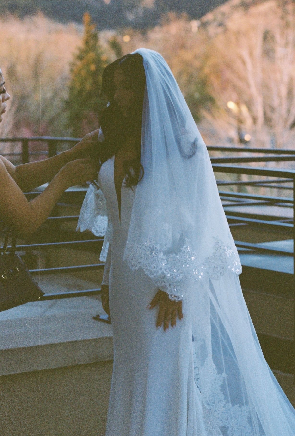 Bridesmaid adjusting cathedral veil with lace trim on bride in white gown on outdoor balcony.