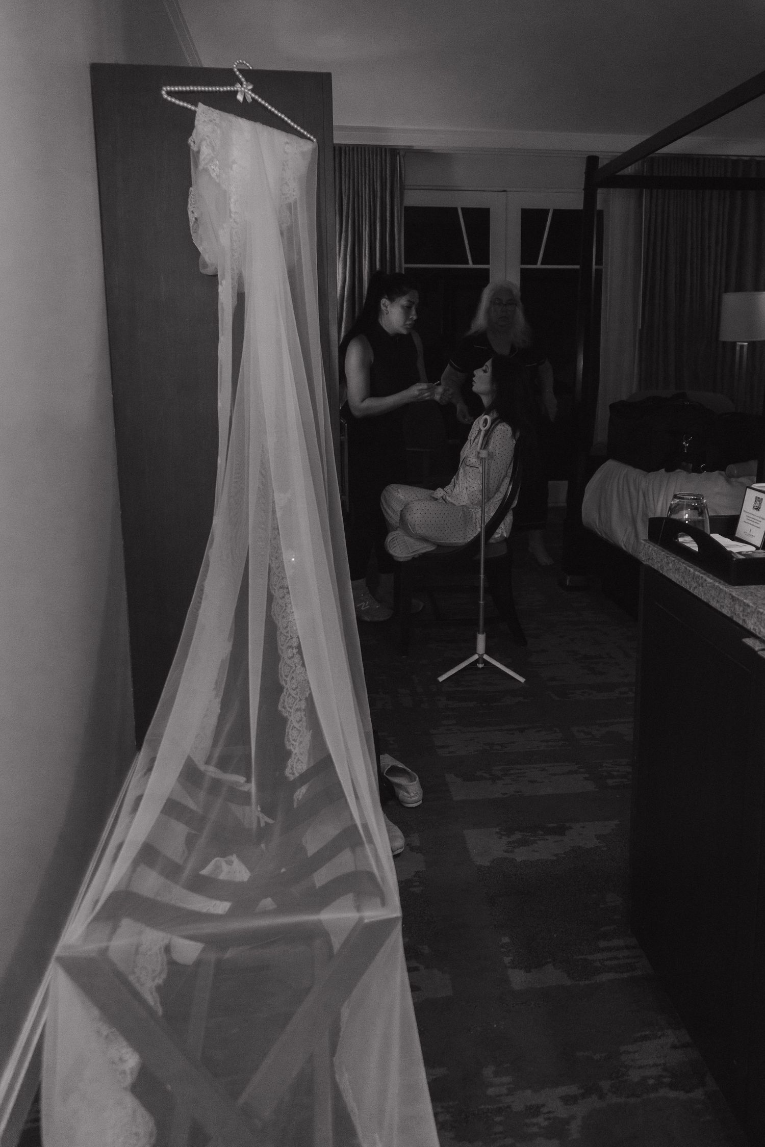 Wedding dress hanging in hotel room while bride gets hair styled in background.