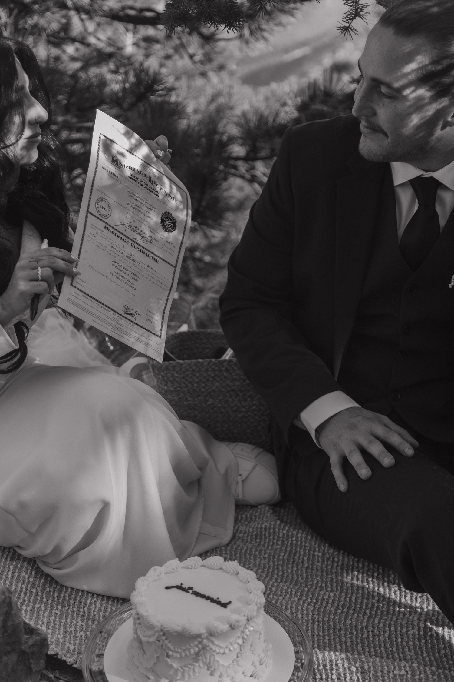 Black and white photo of bride and groom reviewing marriage certificate while seated outdoors with a small wedding cake.