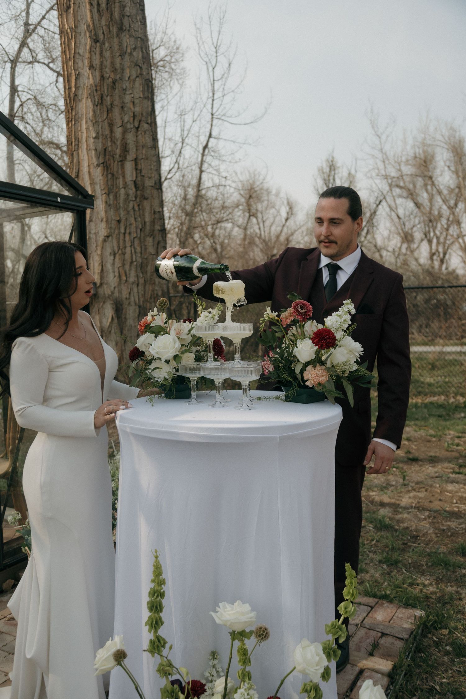 Bride and groom pouring champagne tower at outdoor wedding reception surrounded by white and red floral arrangements.