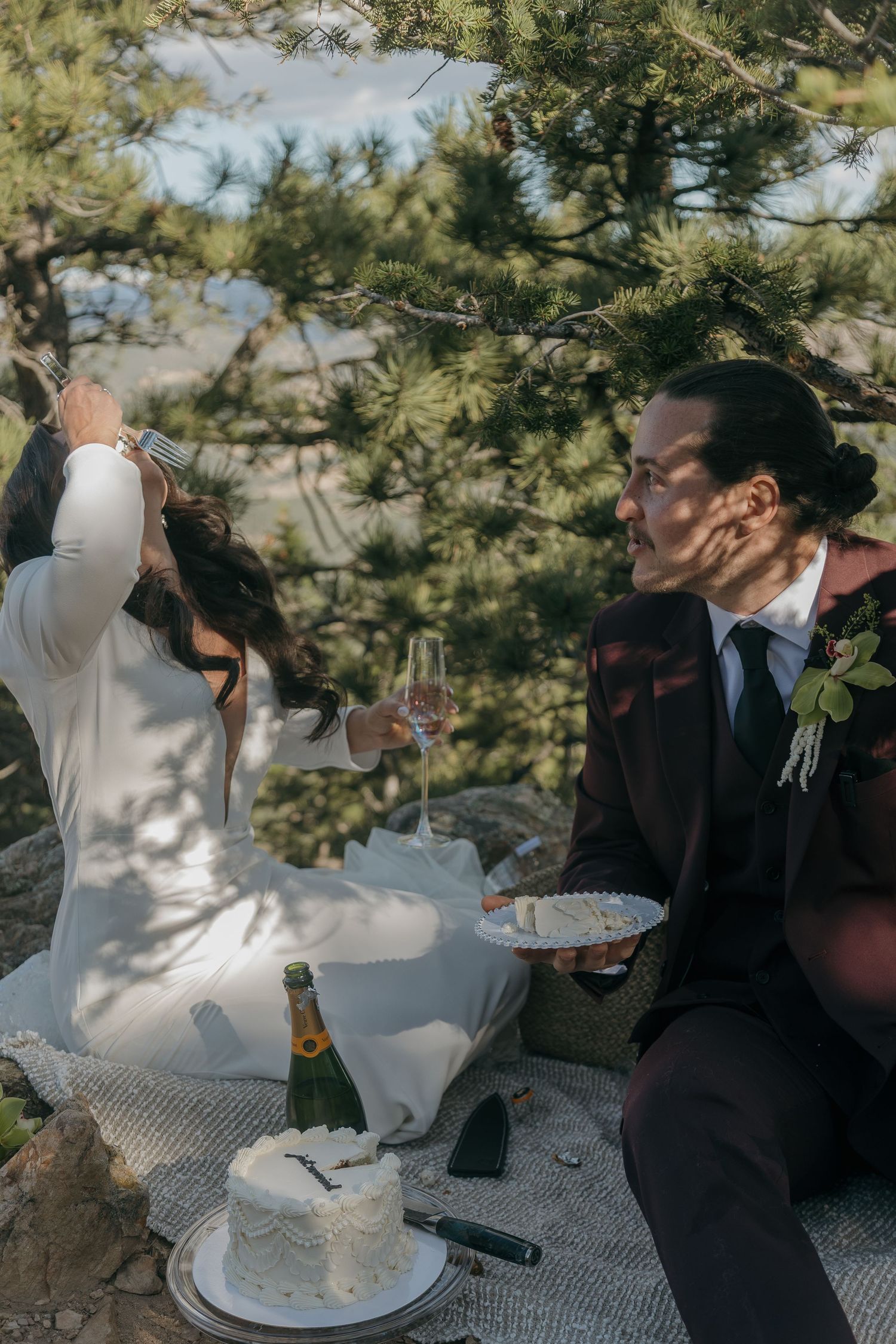 Bride and groom share cake and champagne during an intimate outdoor wedding picnic under pine trees.