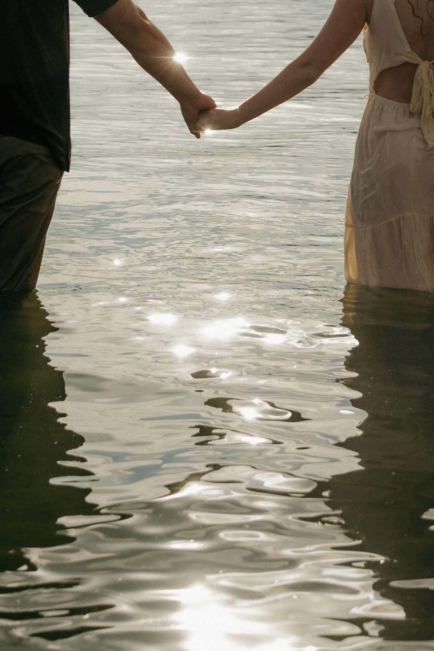 Couple holding hands while standing in sunlit water, reflections sparkling on the surface.
