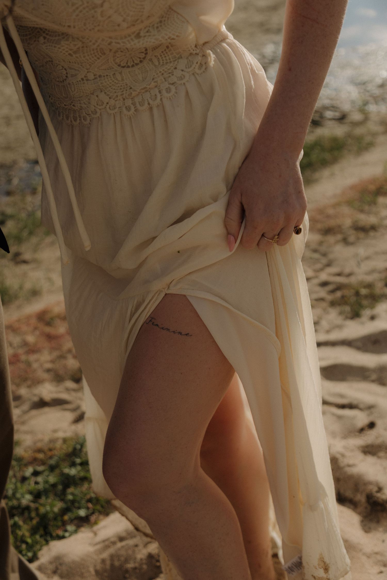 Close-up of woman in cream lace dress lifting hem to reveal thigh tattoo on sandy shore.