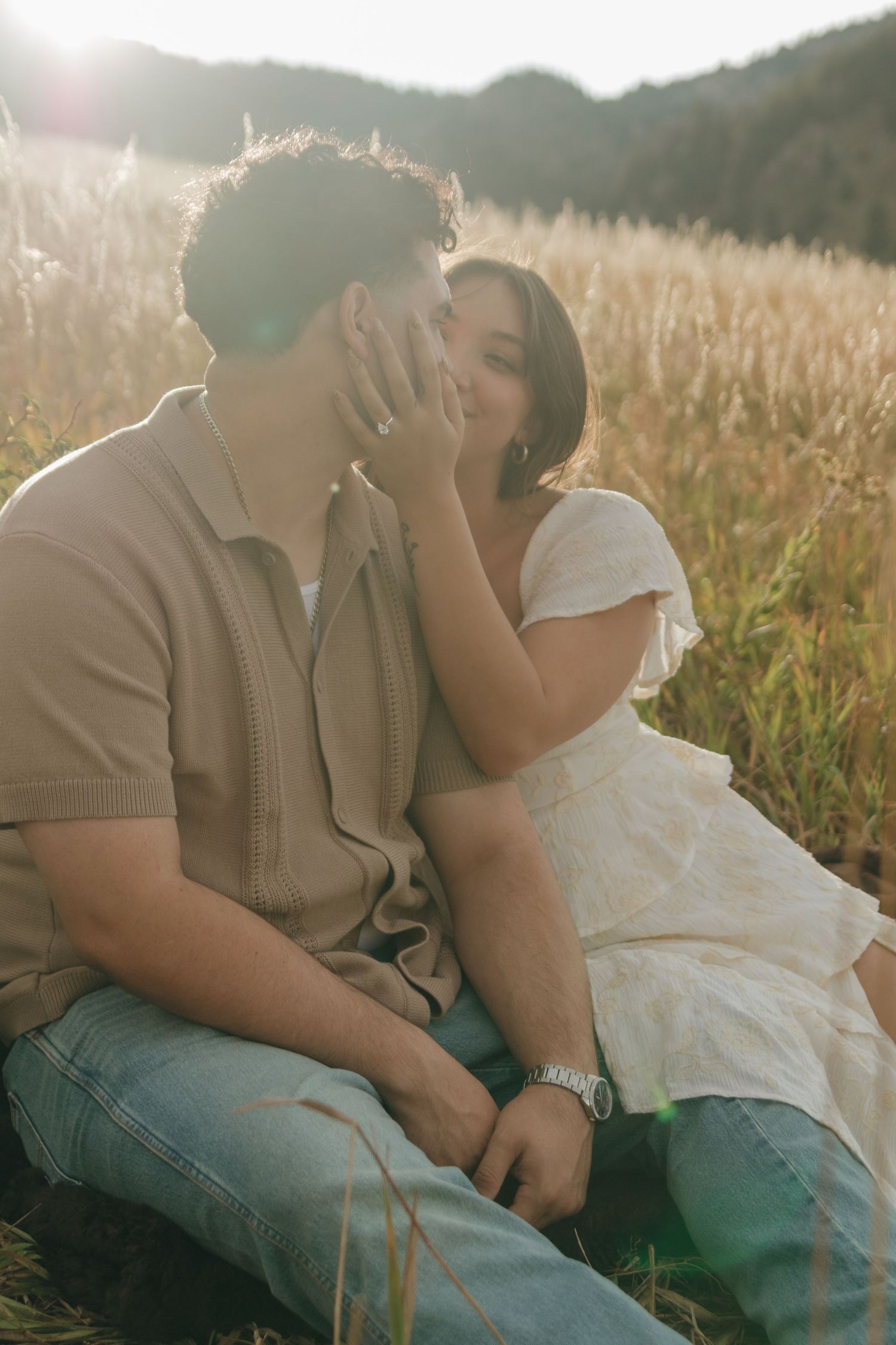 Couple sitting in sun-drenched golden field sharing a tender kiss during a romantic outdoor photo session.