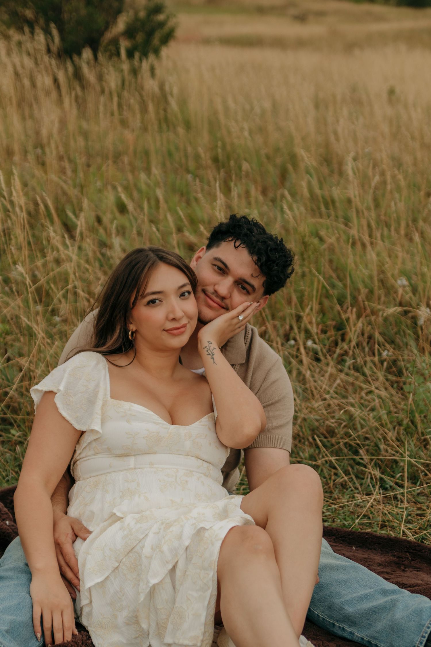 Couple sitting together on a log in a golden meadow, posing for a relaxed outdoor engagement portrait.