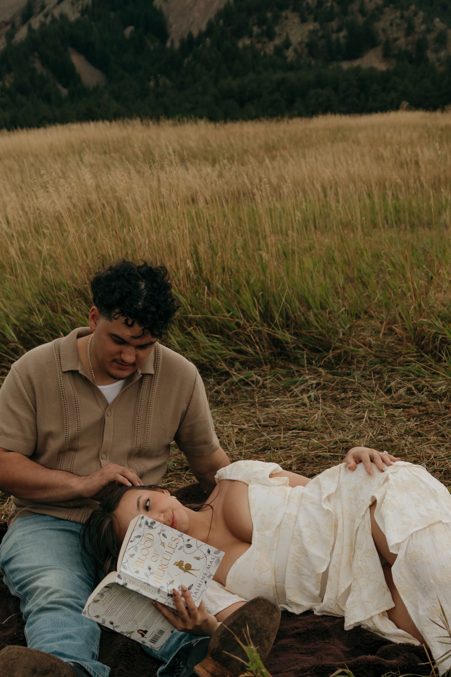 Couple relaxing in field, woman lying down reading book while man sits beside her tenderly.