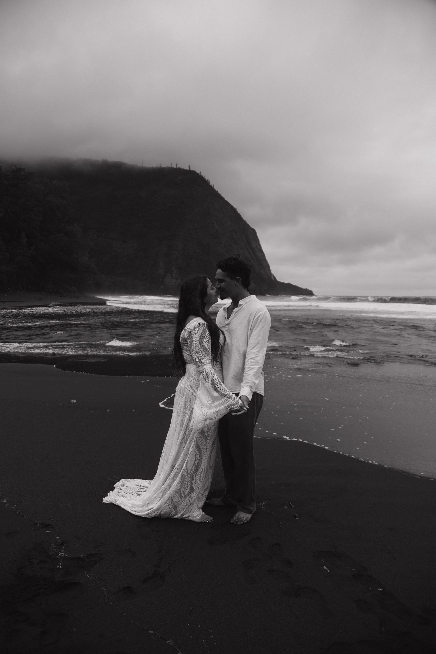 Bride and groom share a kiss on a dramatic black sand beach in black and white photo.