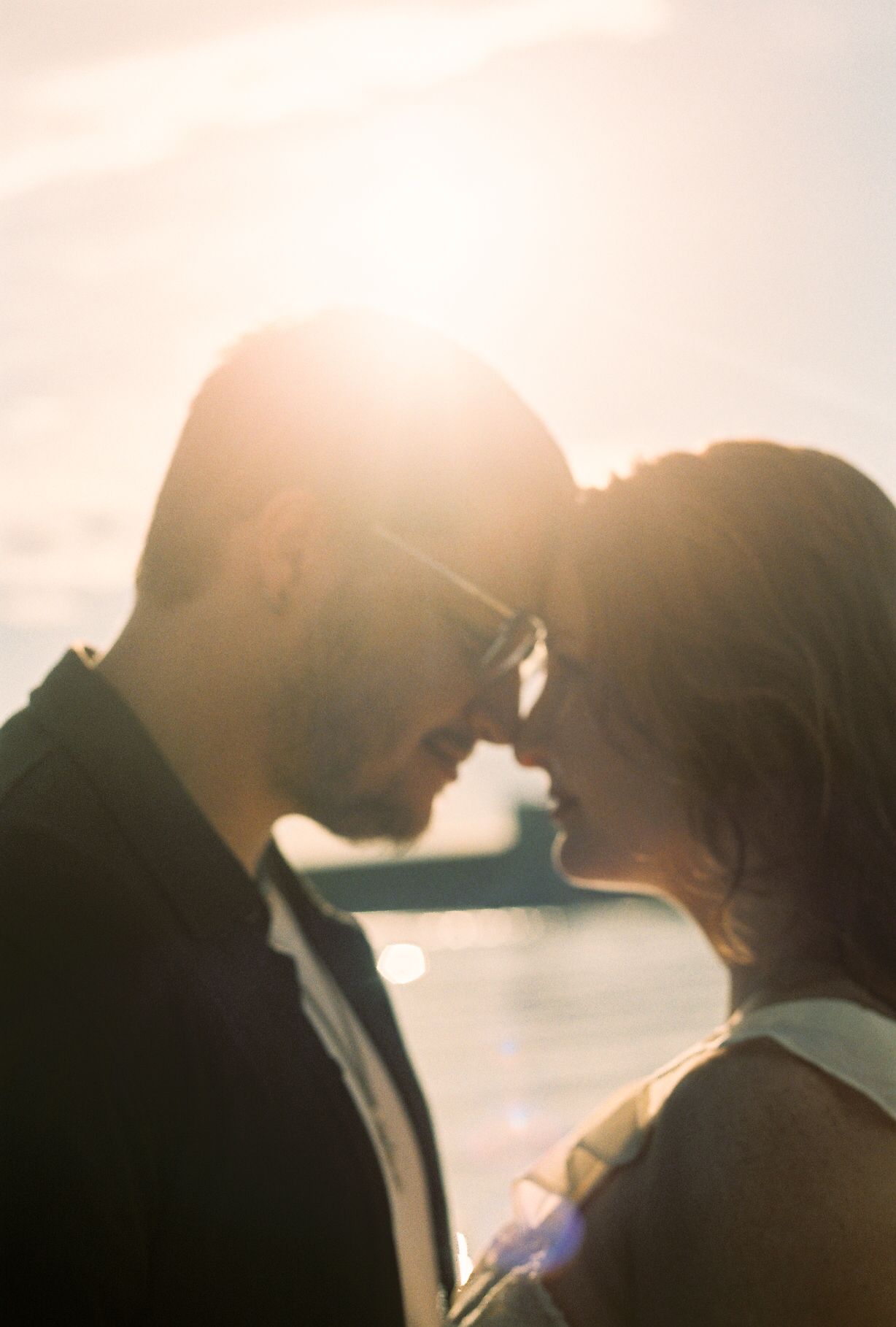 Romantic couple sharing an intimate moment at sunset, foreheads touching with golden backlight glowing behind them.