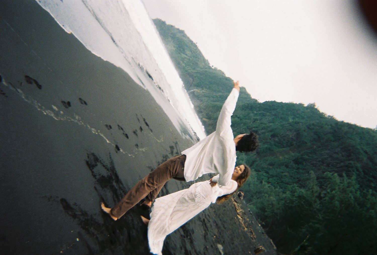 Person in white dress dramatically posed on steep rocky cliff edge with lush green mountains in background.
