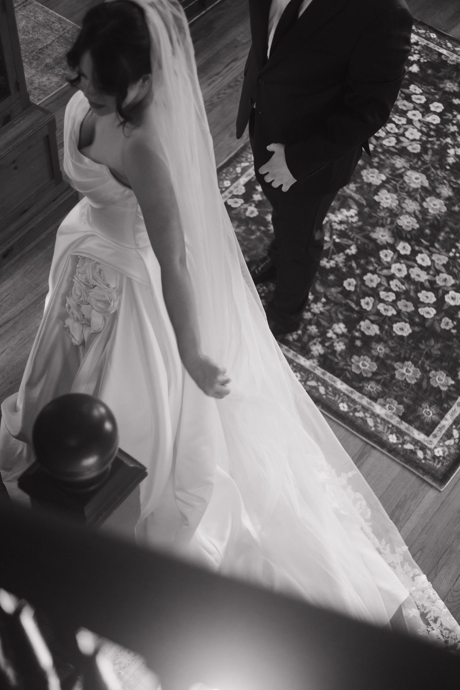 Wedding dress detail shot showing flowing white gown and veil against a patterned carpet.