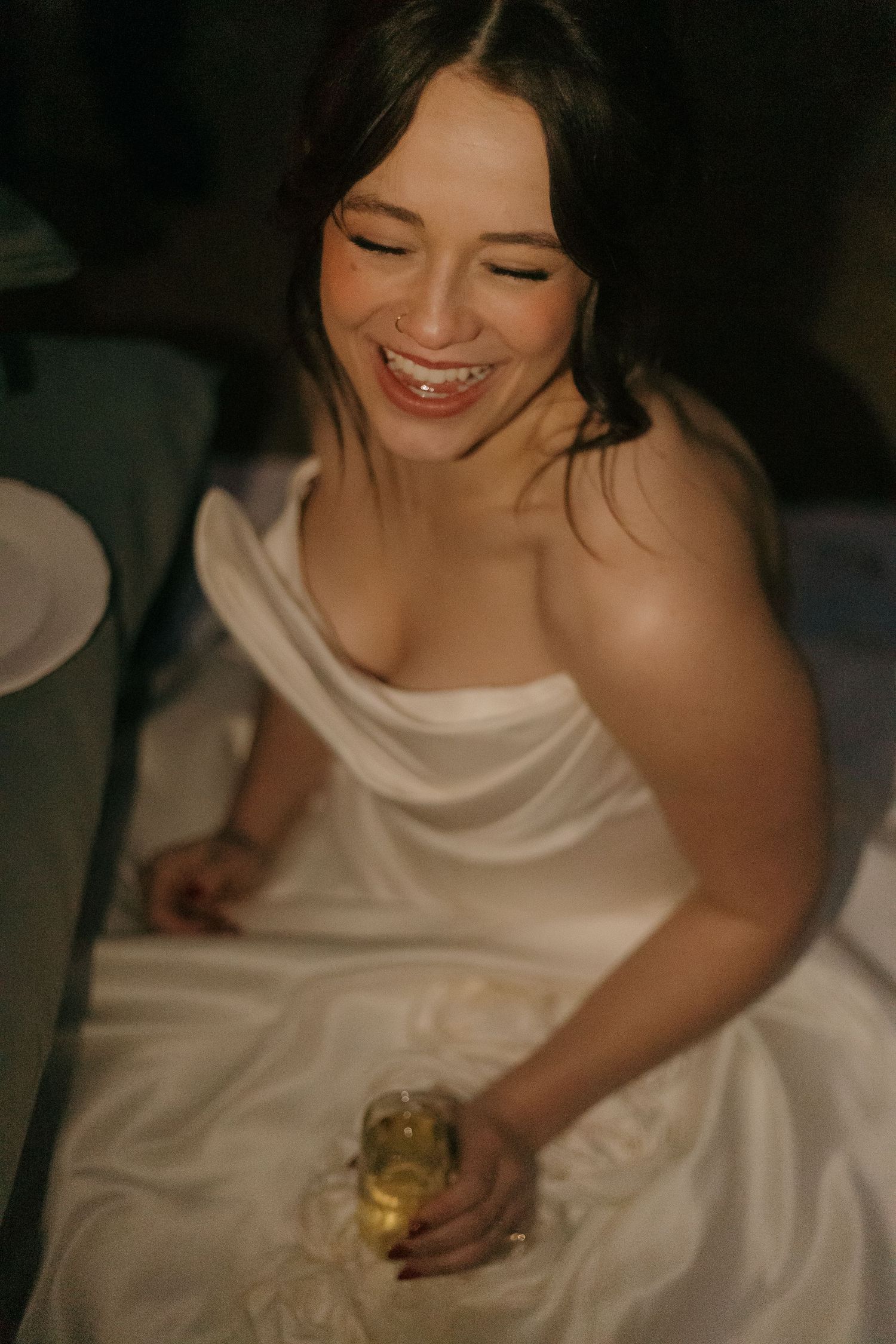 Bride in white dress sitting and holding a drink in a dimly lit setting.