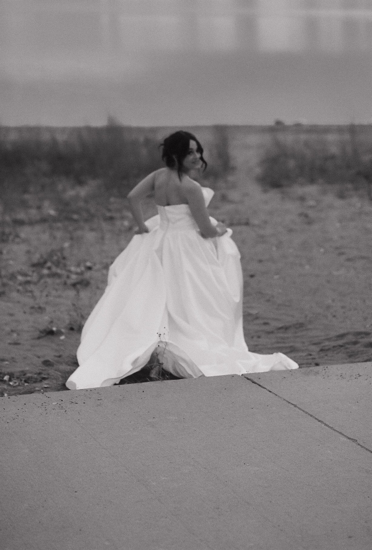Black and white portrait of bride in flowing dress by waterfront.