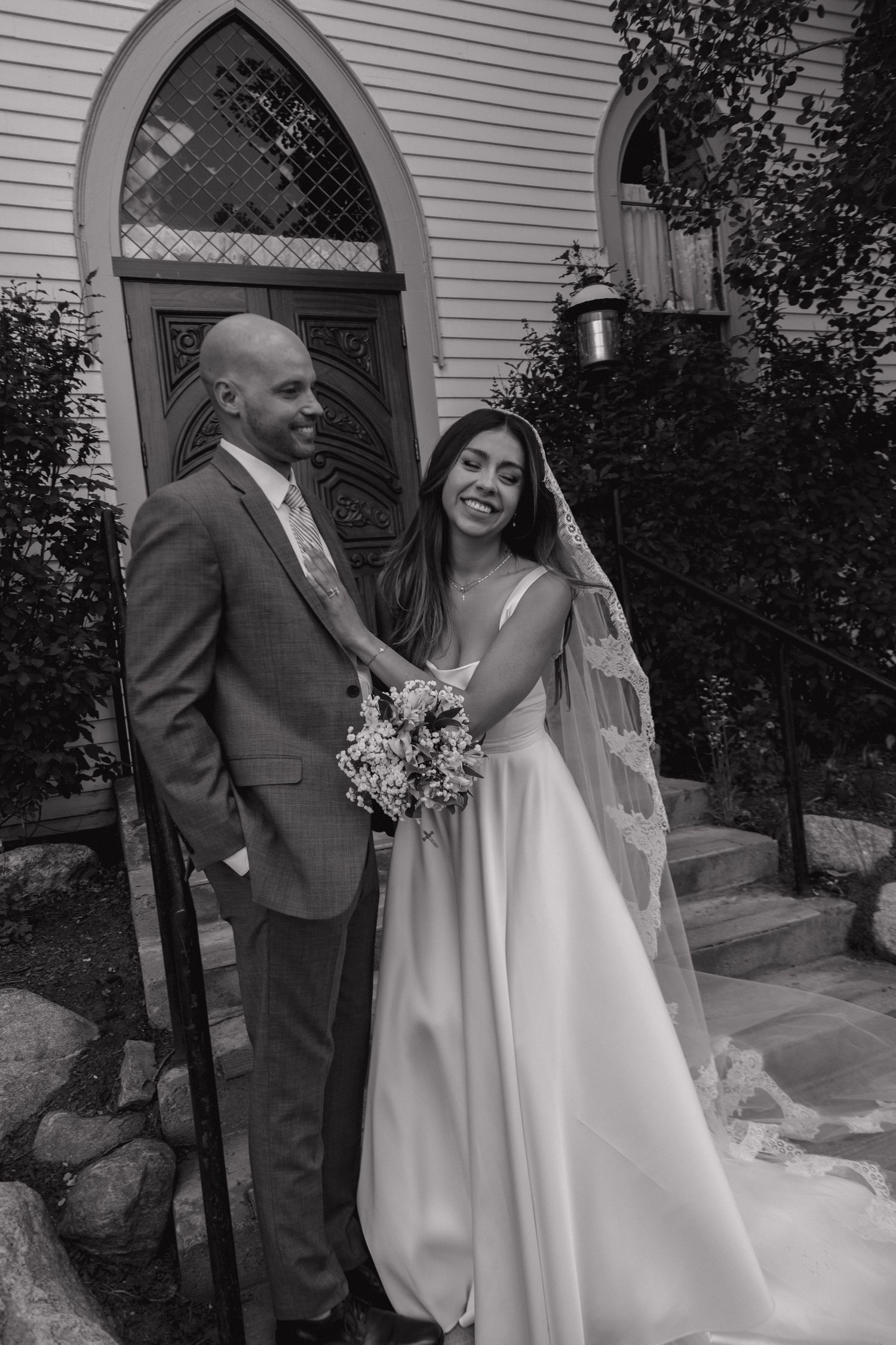 Black and white portrait of smiling bride and groom standing together outside a historic white church after wedding.