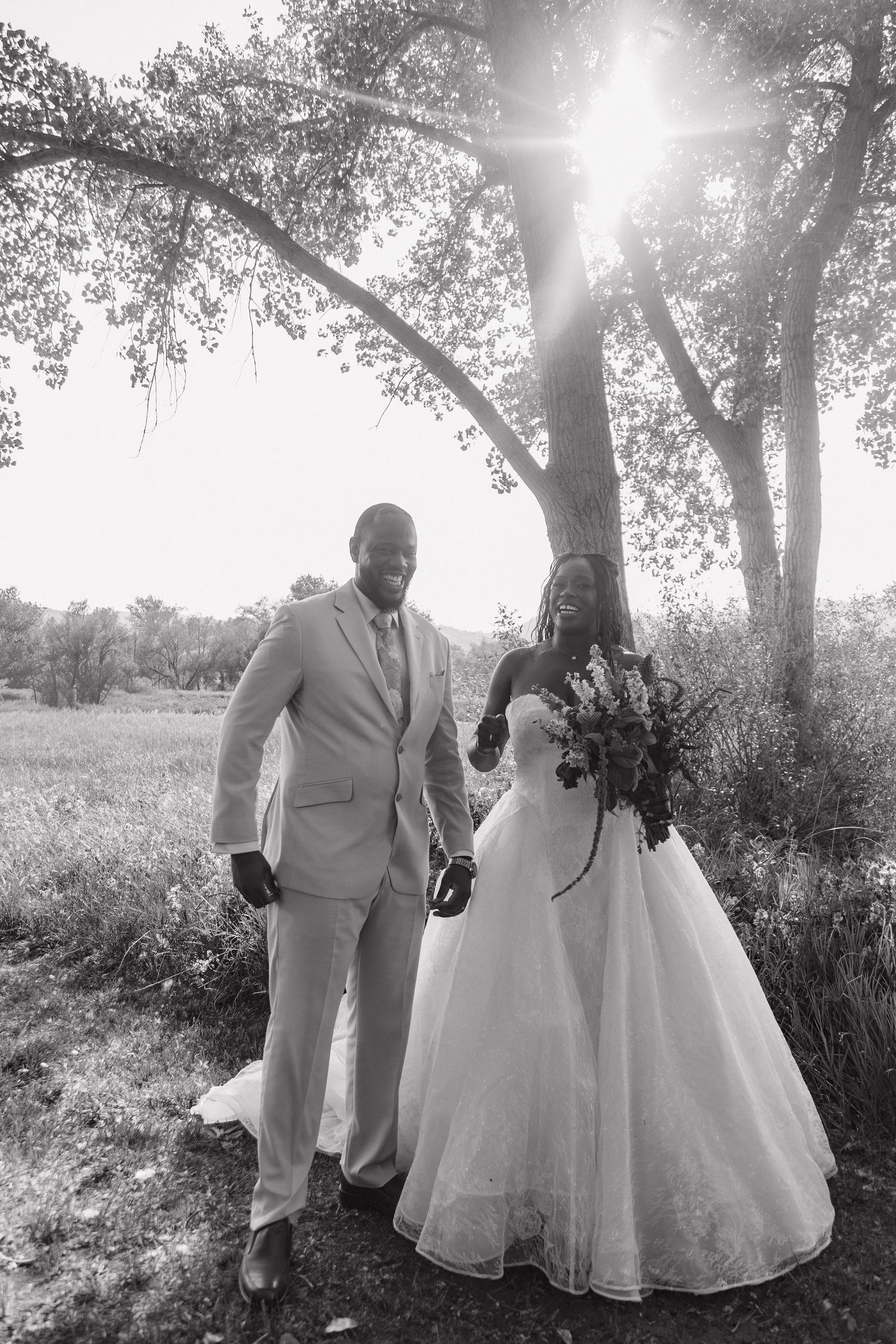 Black and white photo of newlywed couple walking hand in hand under sunlit tree in wedding attire.