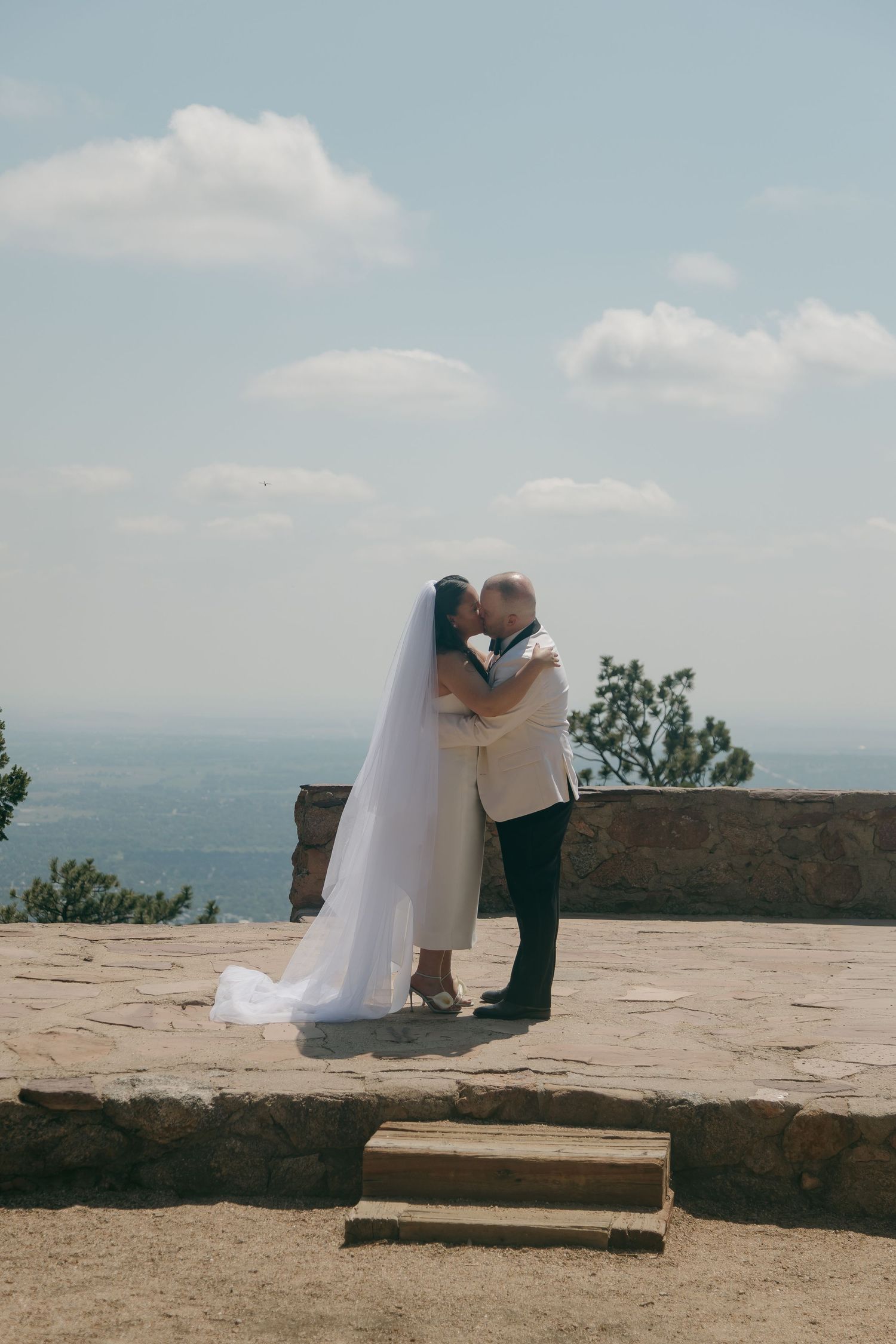 A bride and groom share a romantic kiss on a stone terrace with a scenic sky and landscape behind them.