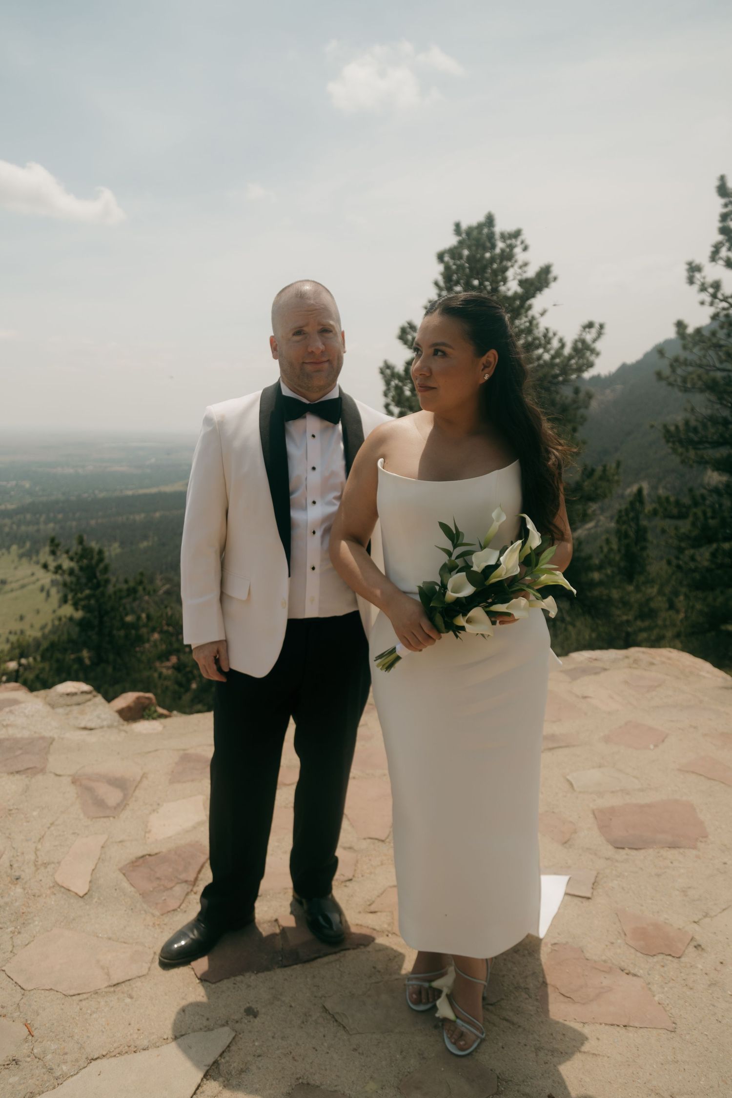 Bride and groom pose with mountain landscape backdrop on sunny wedding day.