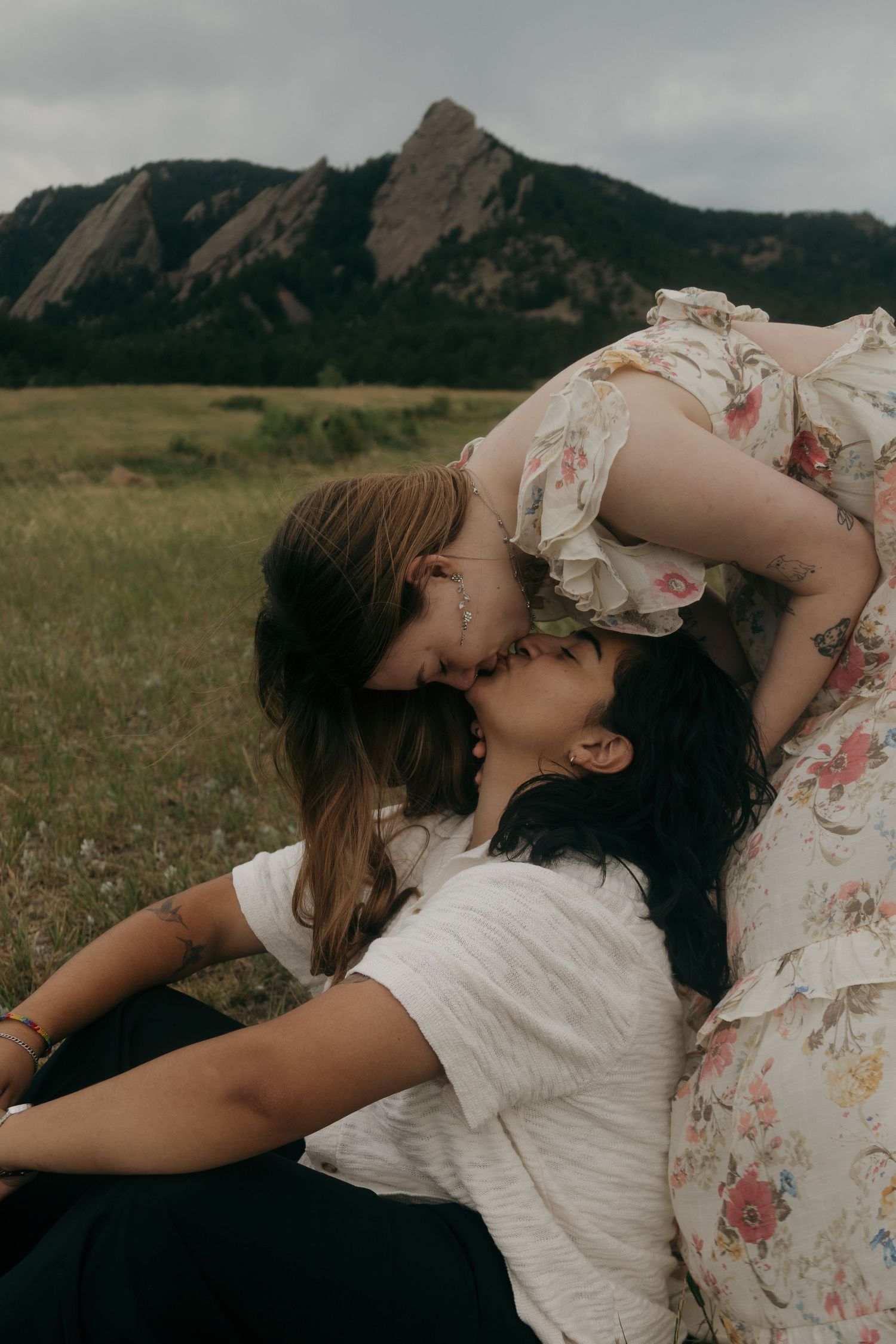 Two women share a kiss in a scenic mountain meadow, one in a floral dress leaning over the other in white.