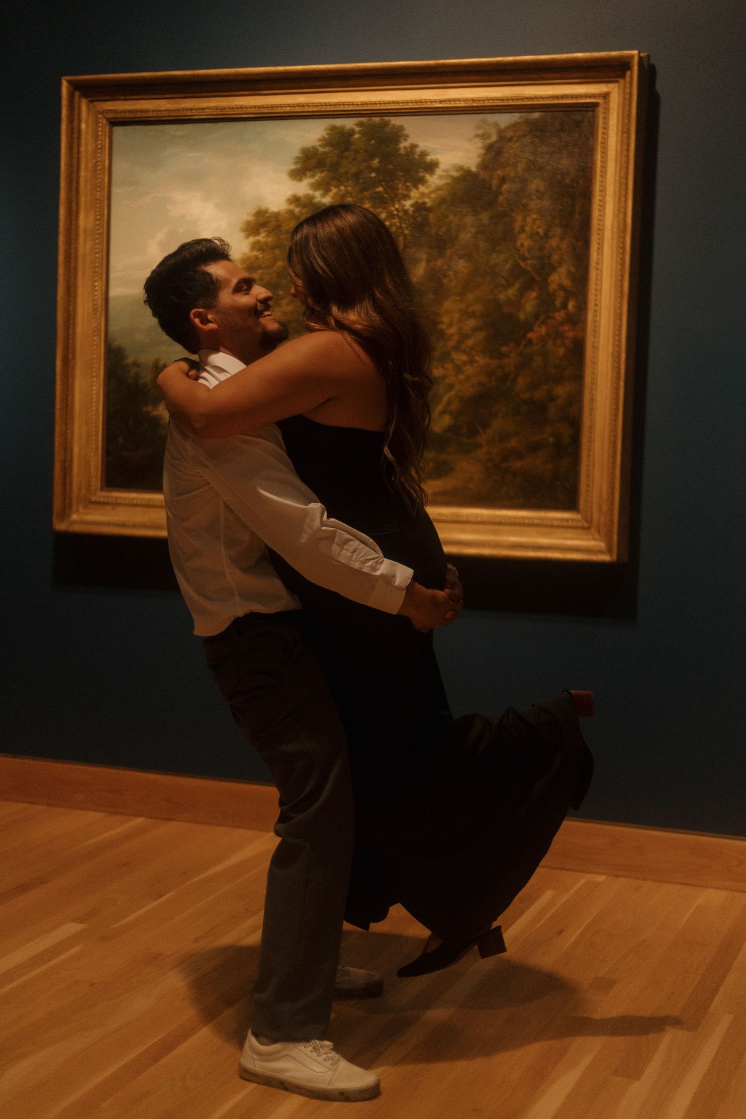 Man lifting woman in black dress as they embrace and kiss before large painting in art museum.