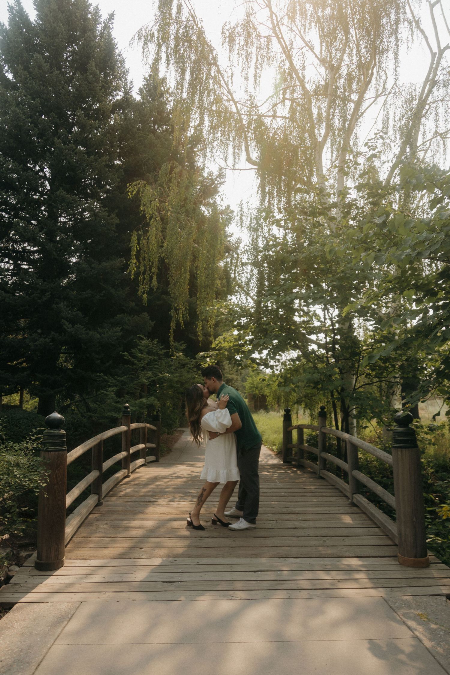 Couple sharing a romantic kiss on a wooden bridge surrounded by lush green trees in a serene park setting.