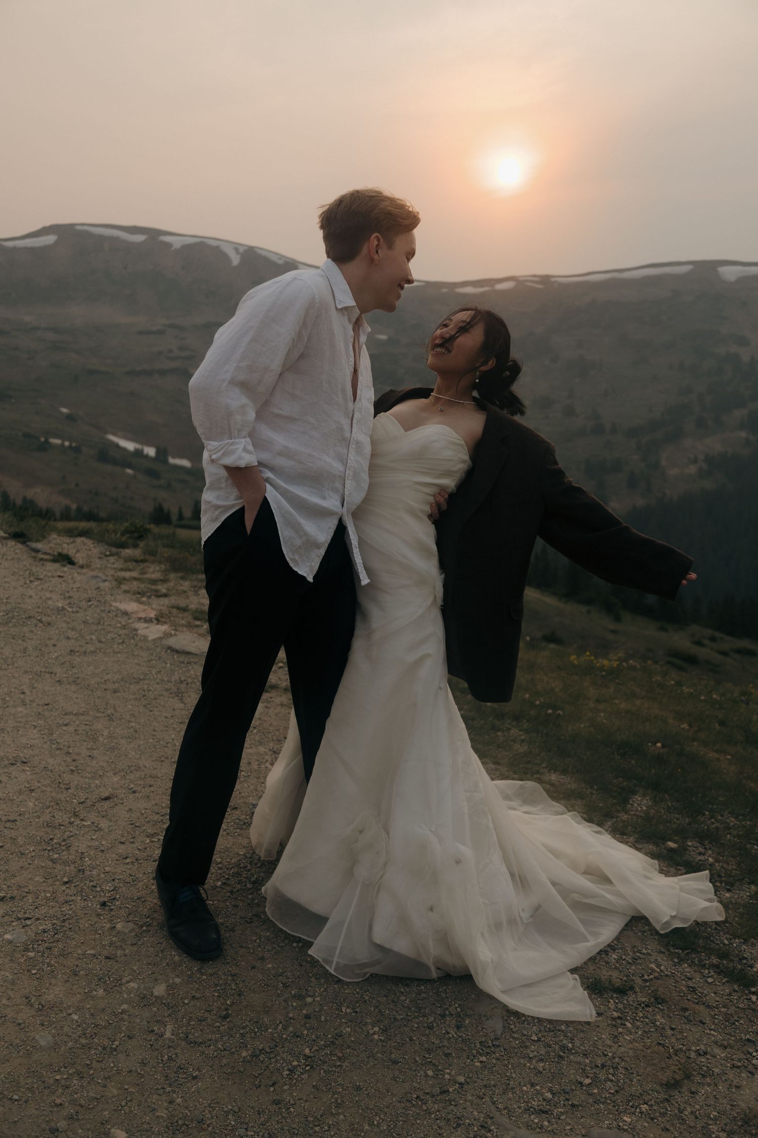 Couple shares a romantic moment at sunset on a mountain overlook, bride in white gown and groom in white shirt.