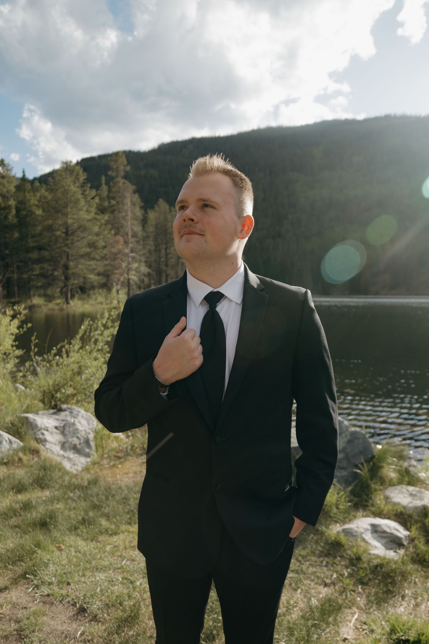 Groom in black suit and tie adjusting jacket lapel outdoors near mountain lake with pine trees and cloudy sky.
