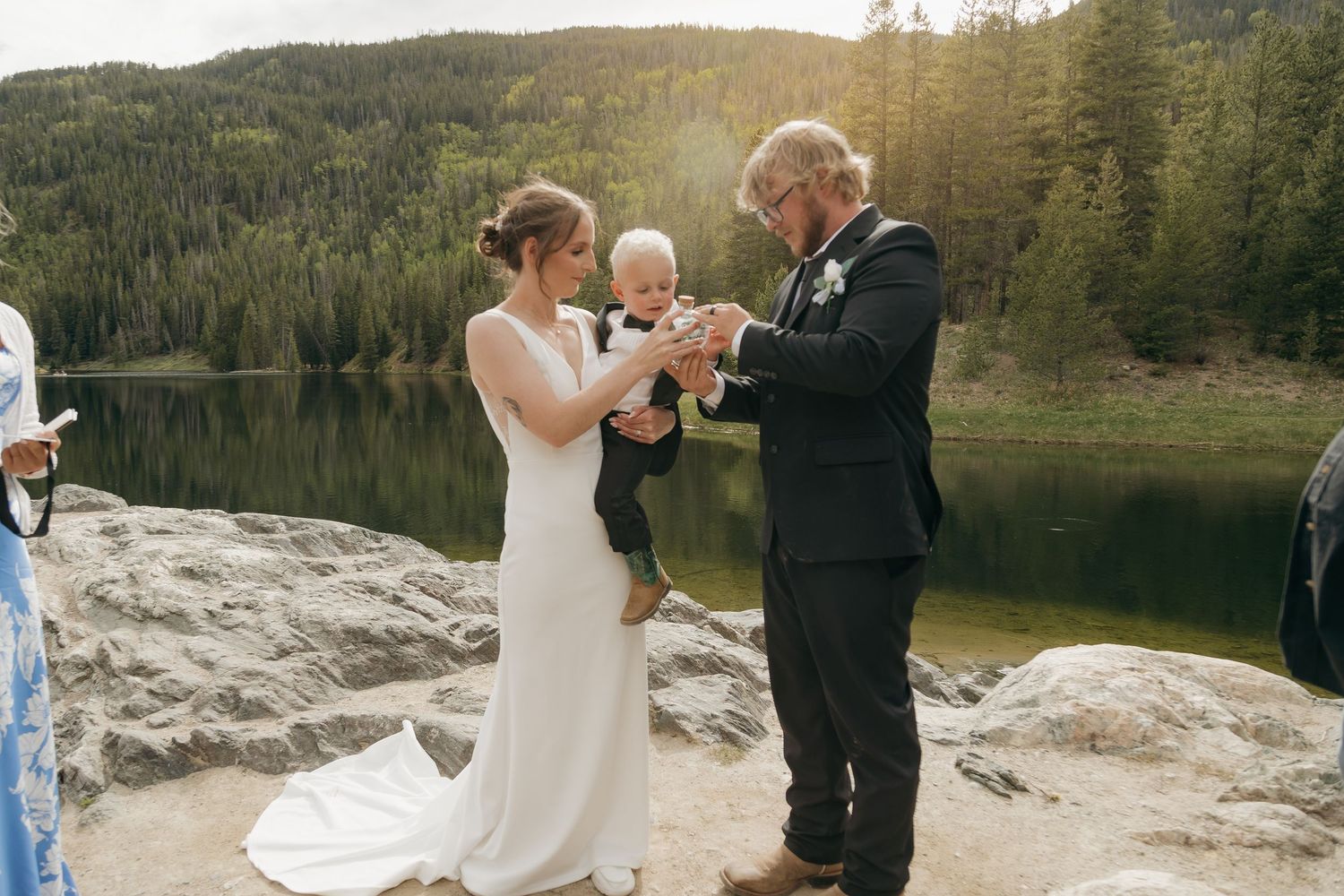 Bride and groom exchange rings outdoors by a mountain lake, holding their baby during an intimate wedding ceremony.