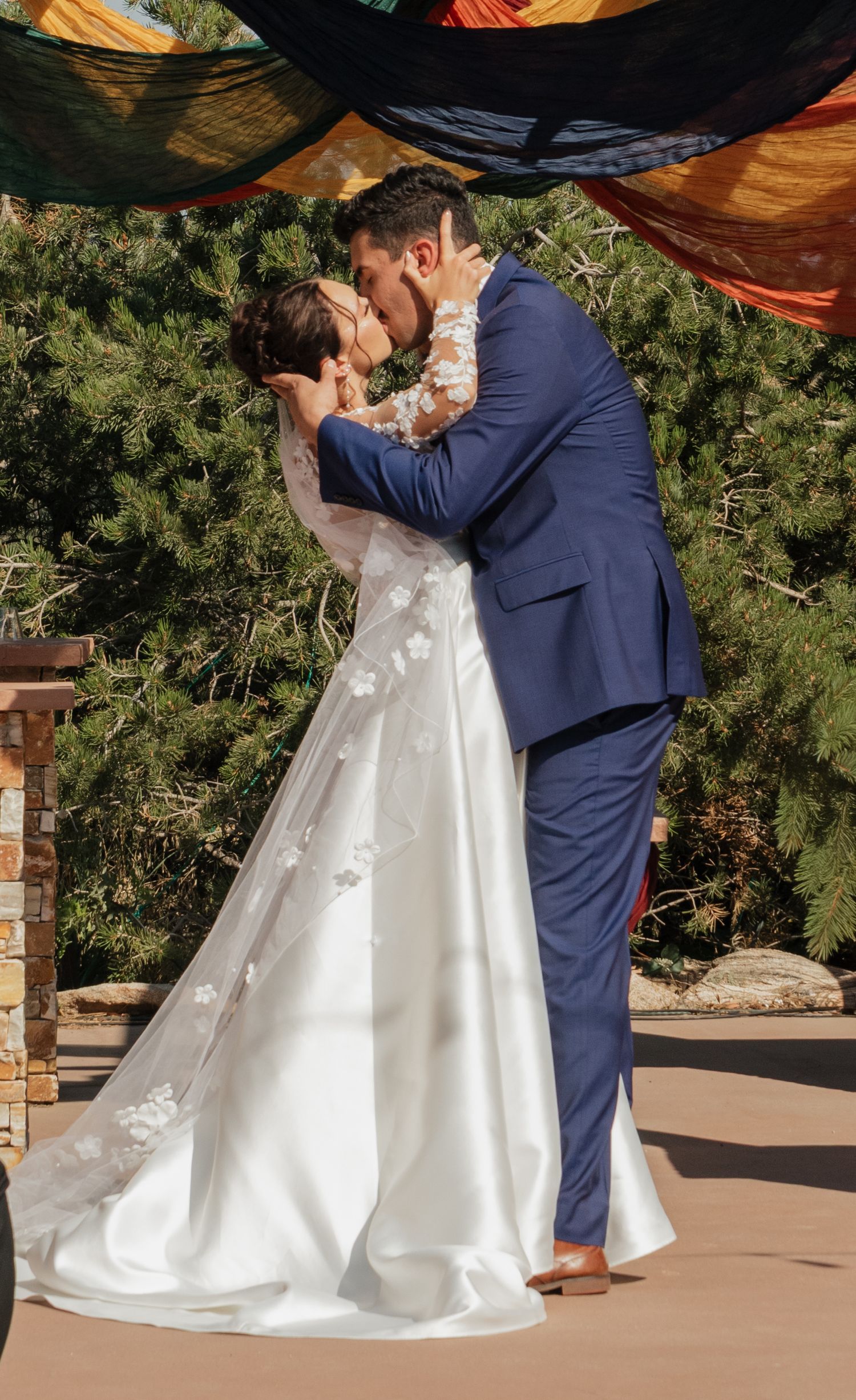 Couple shares their first kiss during outdoor wedding ceremony under decorative canopy.