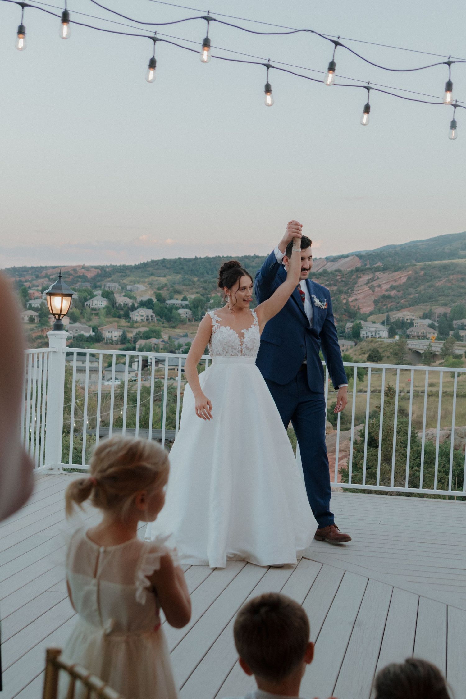 Wedding couple dances on outdoor deck with string lights and mountain views at dusk.