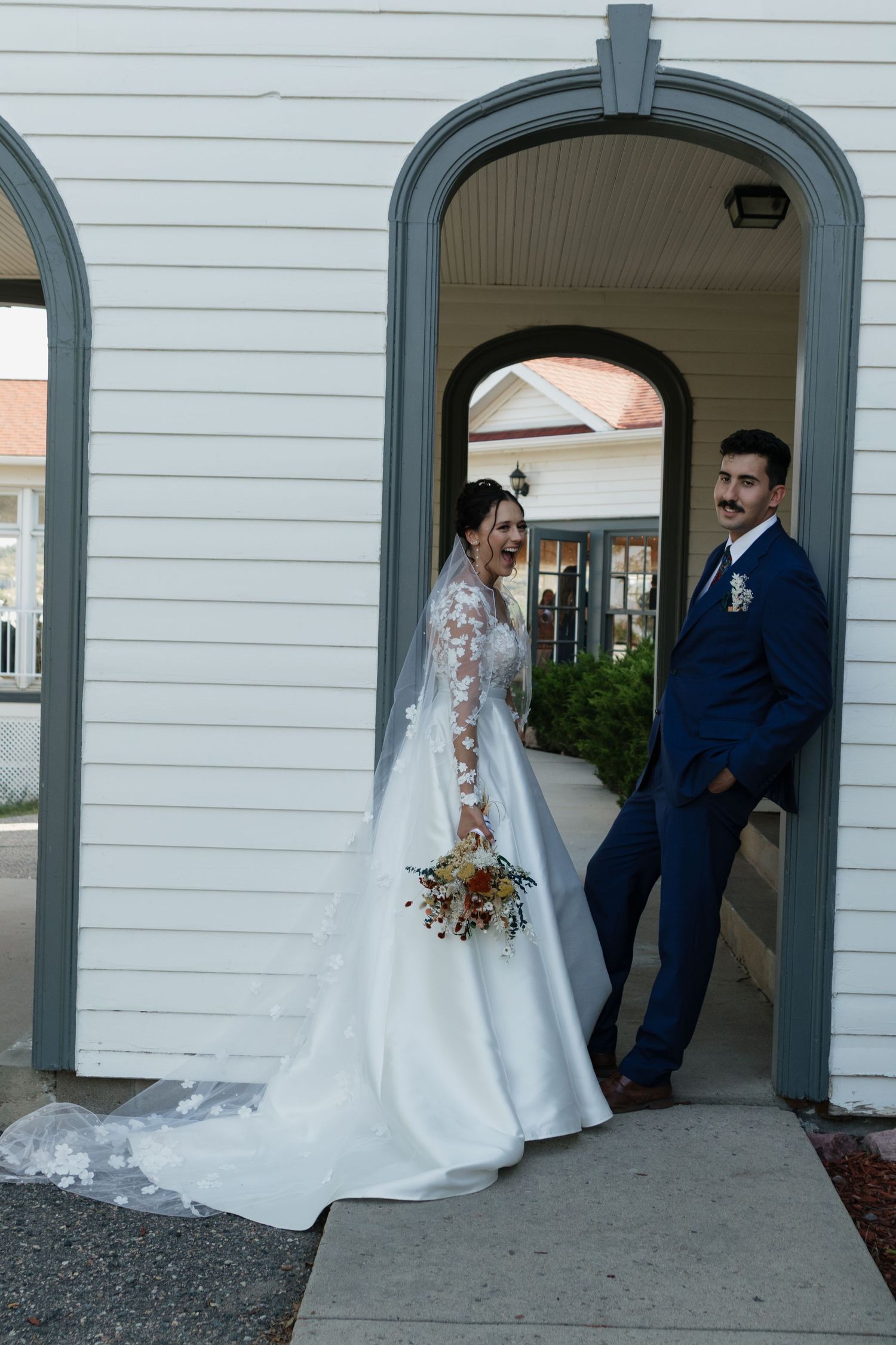 A couple poses in formal wedding attire under white arched doorways of a traditional building.