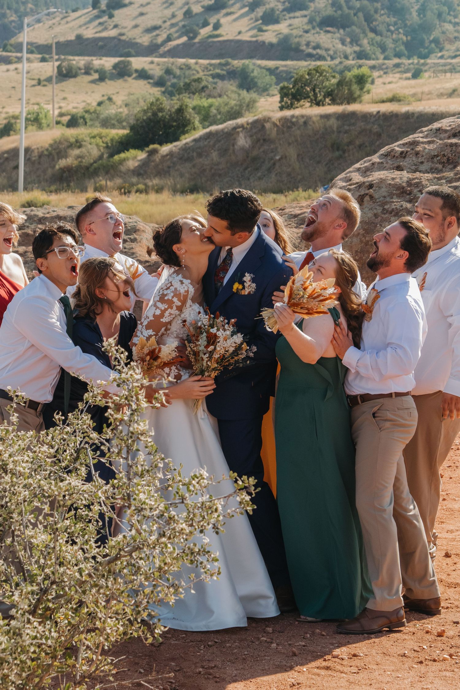 Wedding guests gather for group photo in rustic outdoor setting with mountains in background.