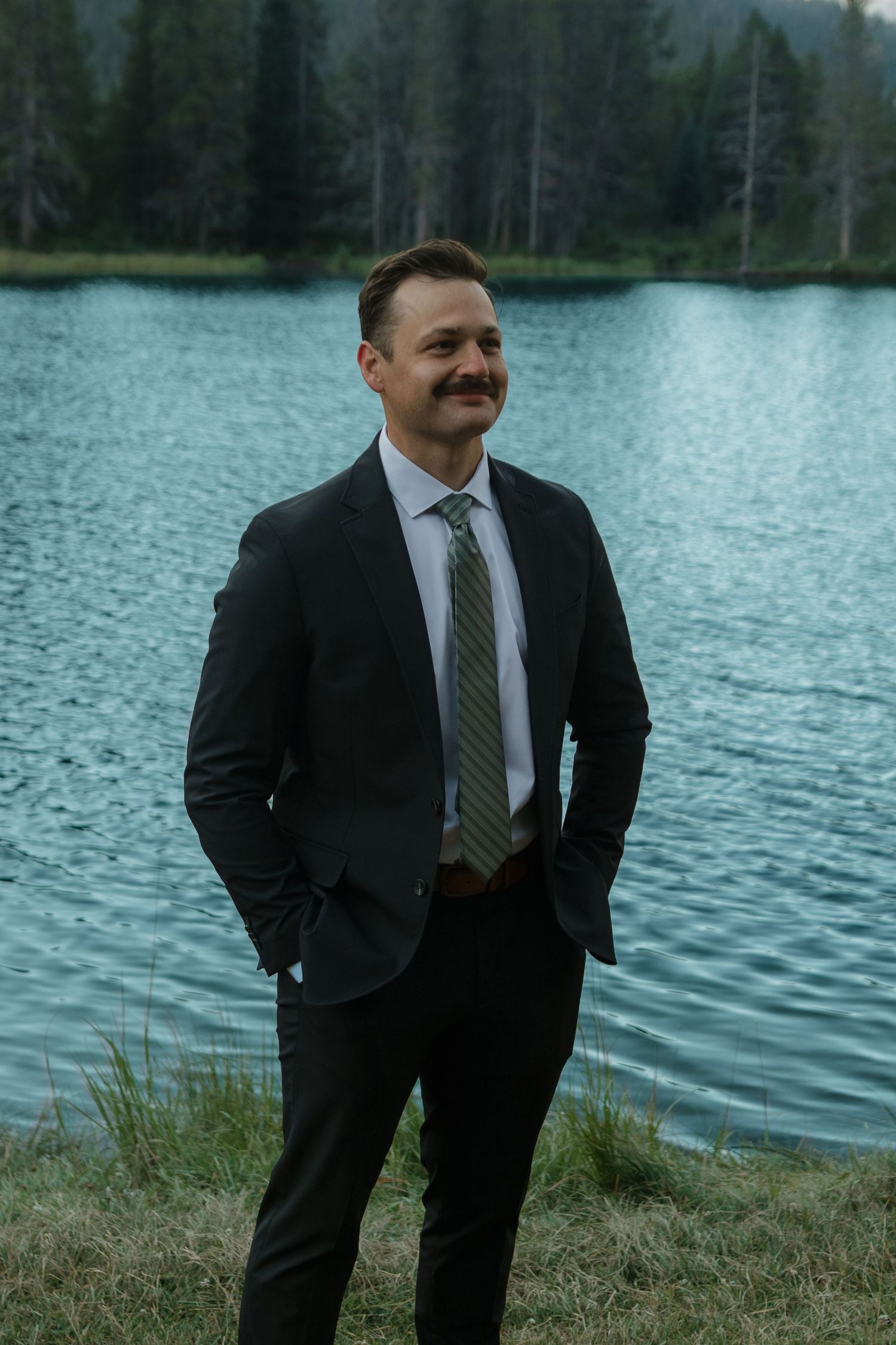 Suited man stands by scenic mountain lake with pine trees in background.