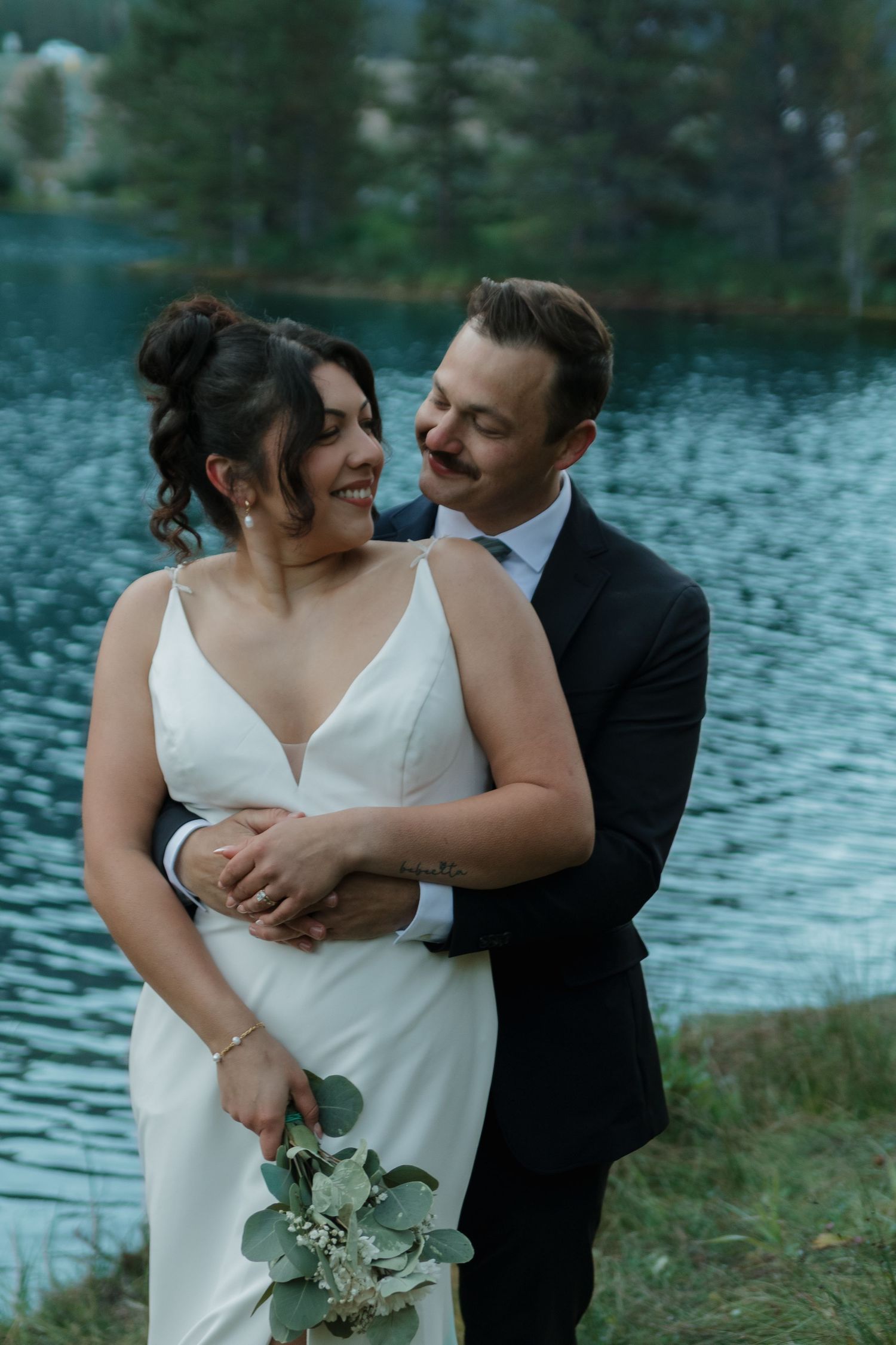 Romantic wedding couple embraces by tranquil lakeside with lush greenery and rippling water in background.