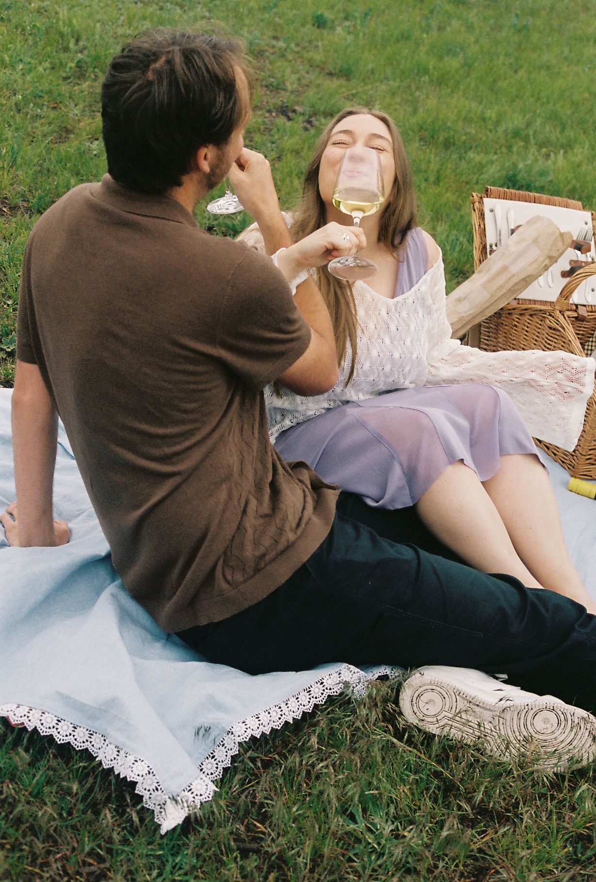 Couple enjoying a romantic outdoor picnic on a blanket, toasting with white wine glasses in a green field.