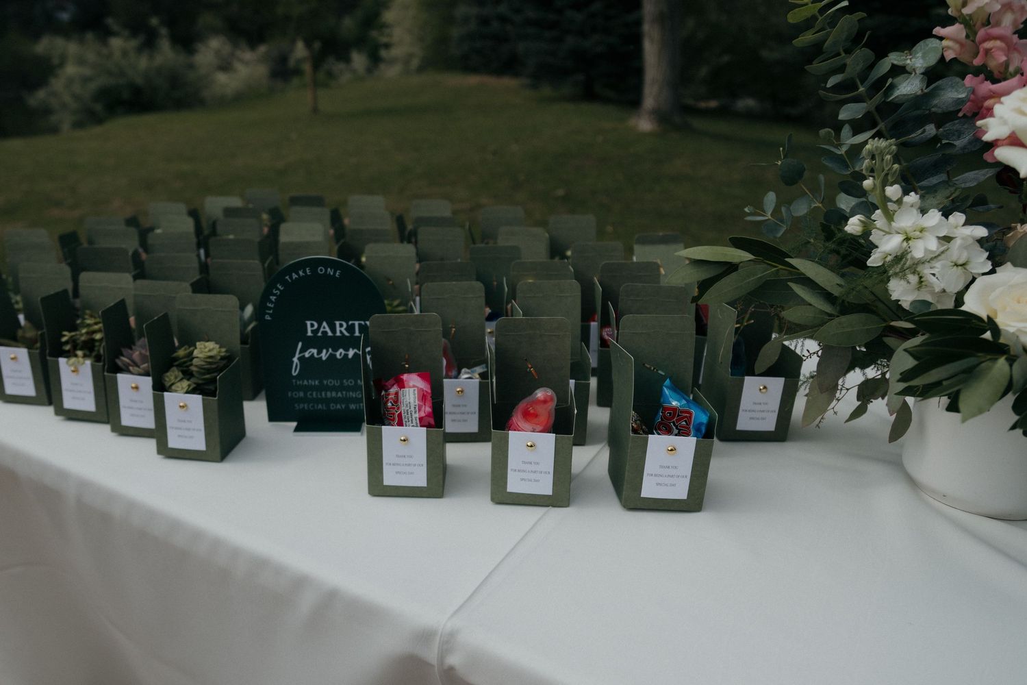 Dark green wedding party favor boxes arranged on a white table with floral decorations and a party favors sign.