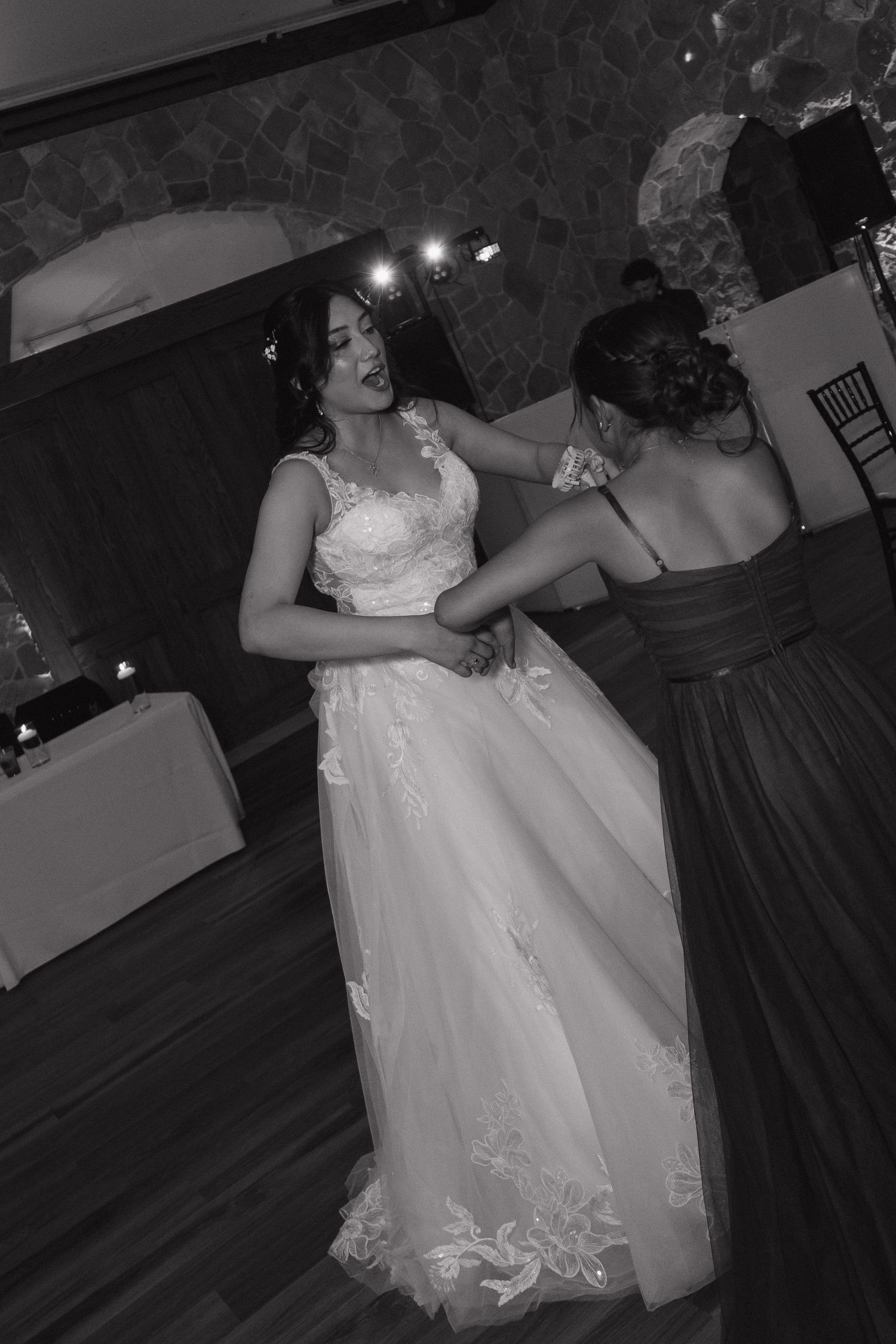 Bride dancing with bridesmaid in black dress at wedding reception in black and white.
