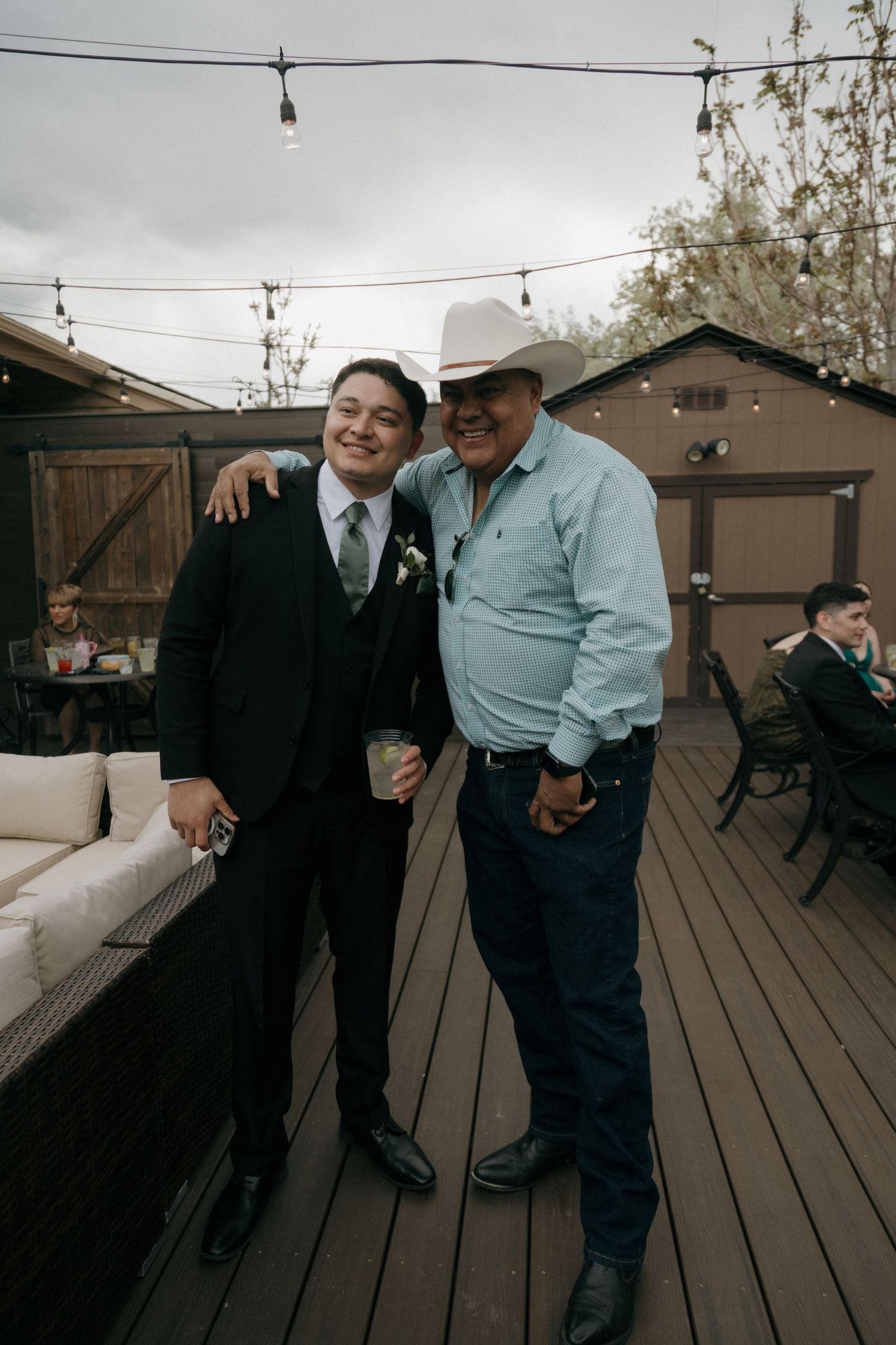 Groom in black suit poses with older man in cowboy hat on outdoor deck.