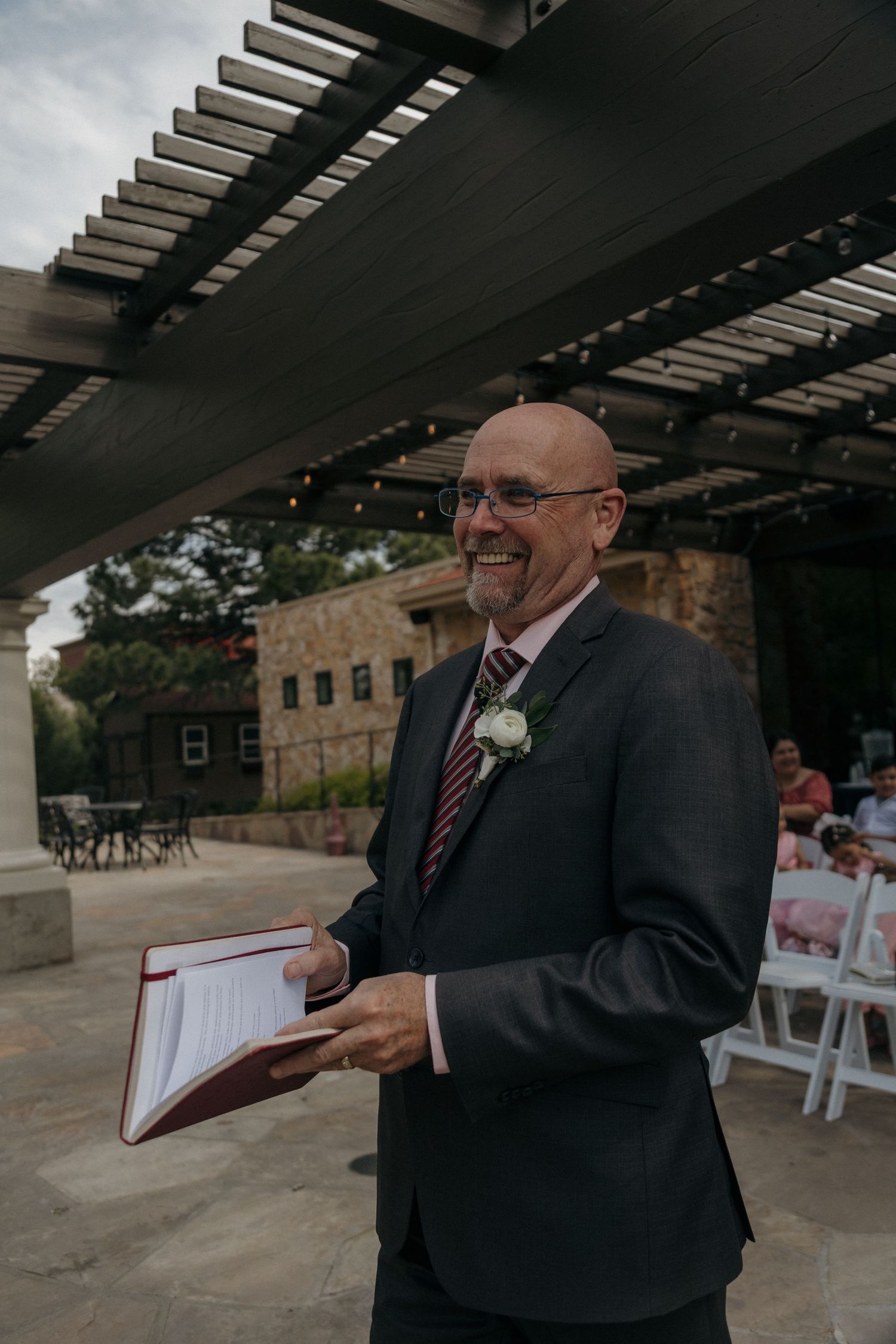 Smiling bald officiant in suit holding ceremony book at an outdoor rustic stone wedding venue.