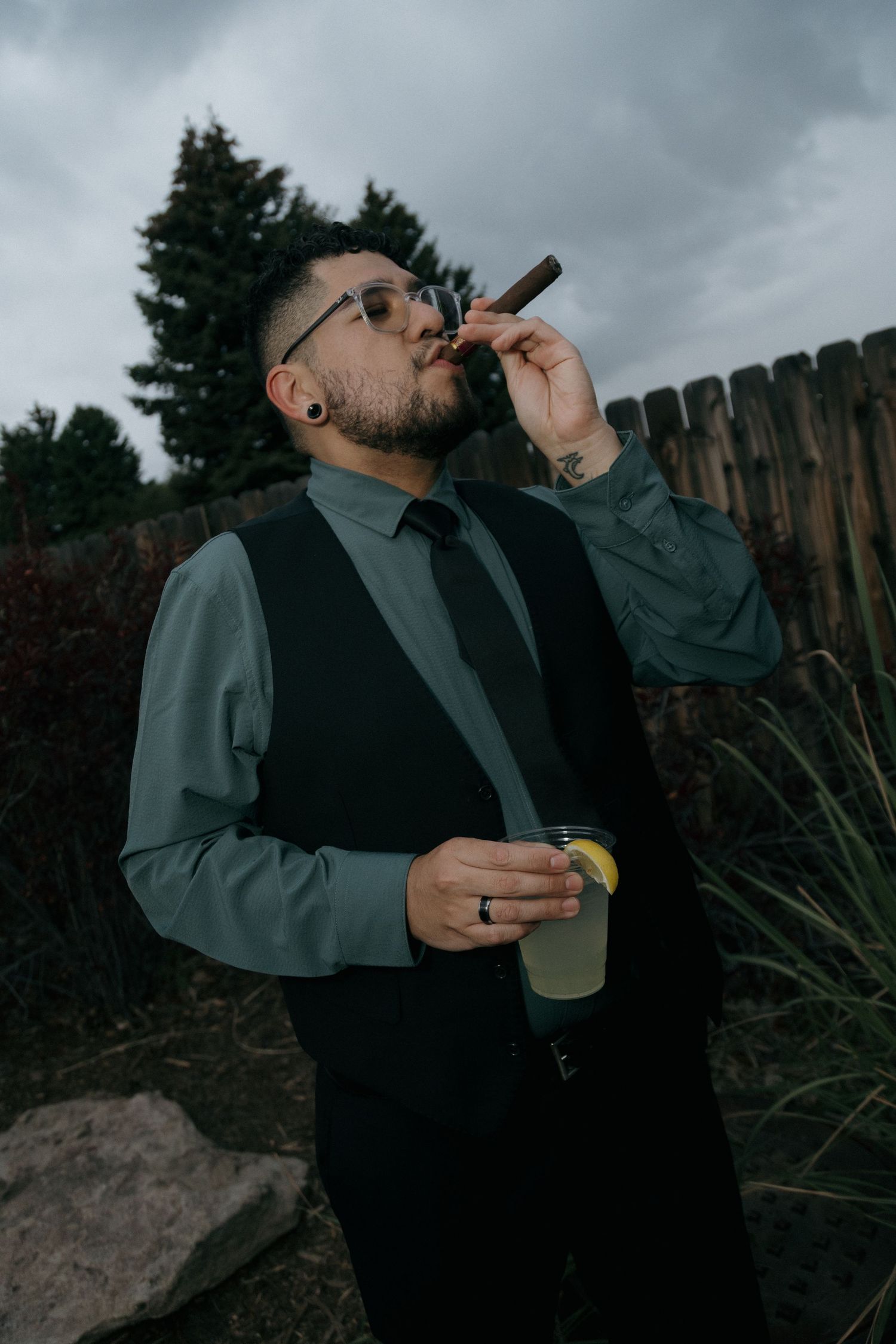 Stylish groomsman in black vest smokes cigar and holds cocktail outdoors under cloudy sky.