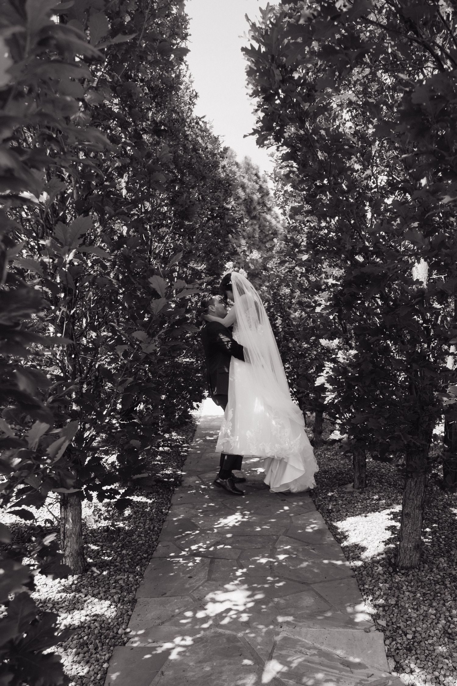 Black and white photo of a bride and groom sharing a kiss on a tree-lined garden pathway.