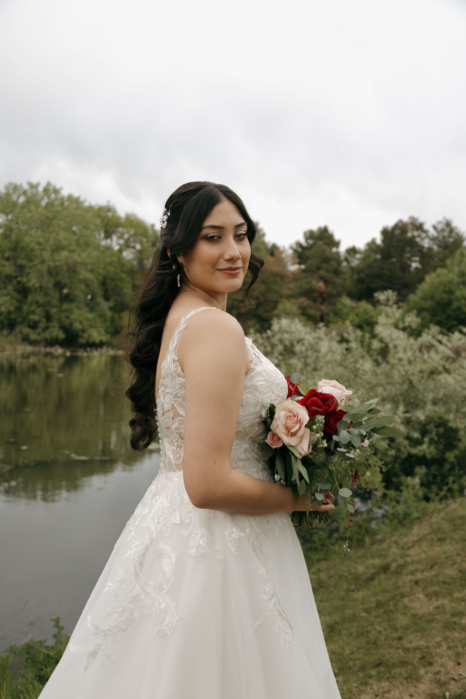 Elegant bride in lace wedding gown holding red and pink bouquet beside serene garden pond.
