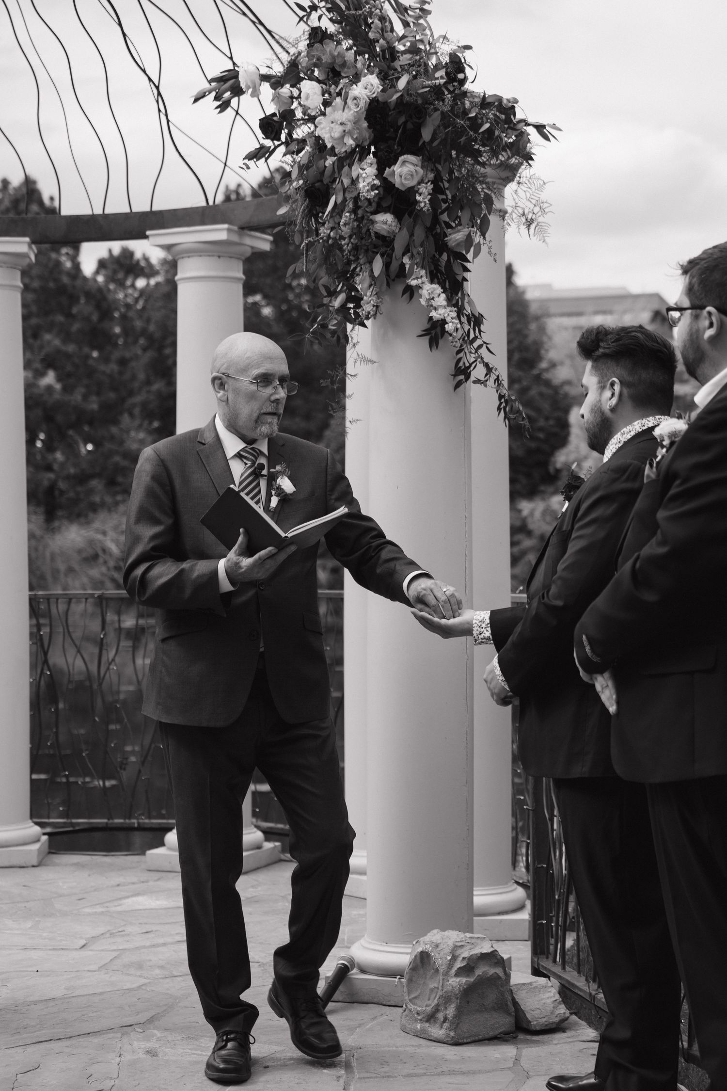Officiant gestures during outdoor wedding ceremony at floral arch with groom and guest in black and white.