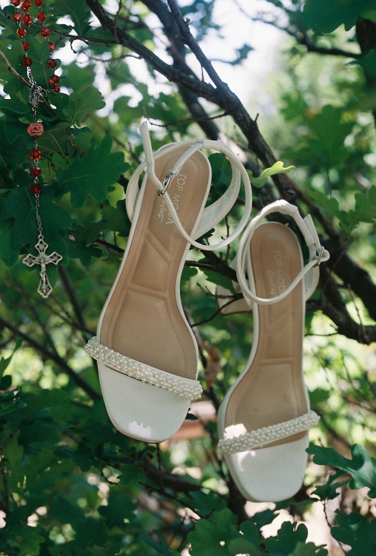 White bridal sandals with pearl straps hanging from a tree branch beside a cross rosary.