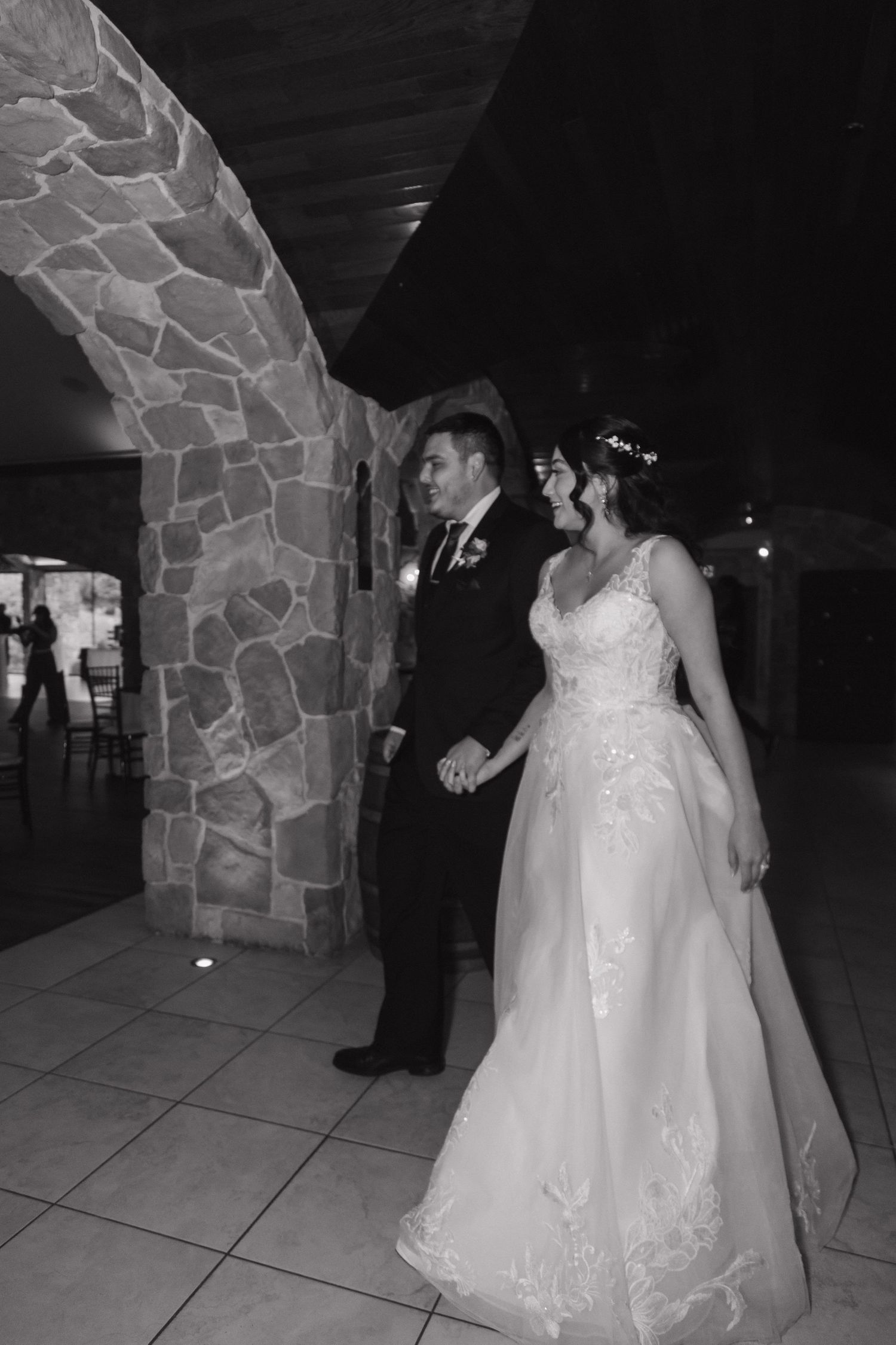 Black and white photo of bride and groom holding hands and smiling inside a rustic stone arch wedding venue.