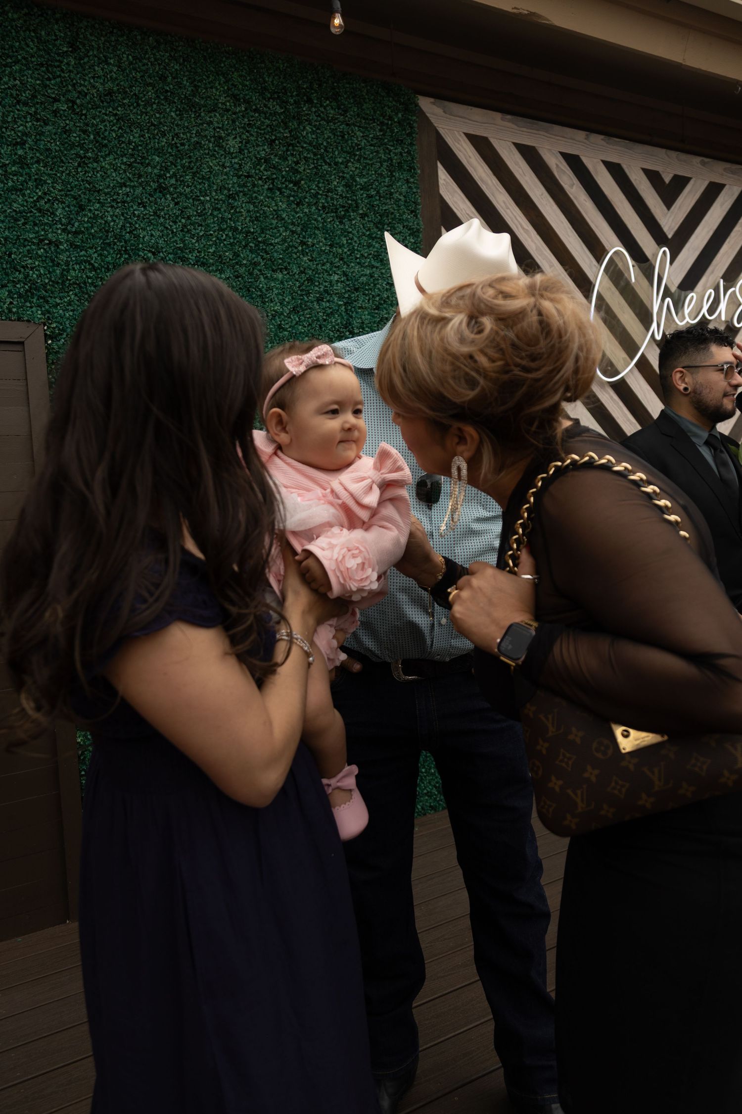 Elderly woman admires an adorable baby in pink at a wedding reception near a green hedge backdrop.
