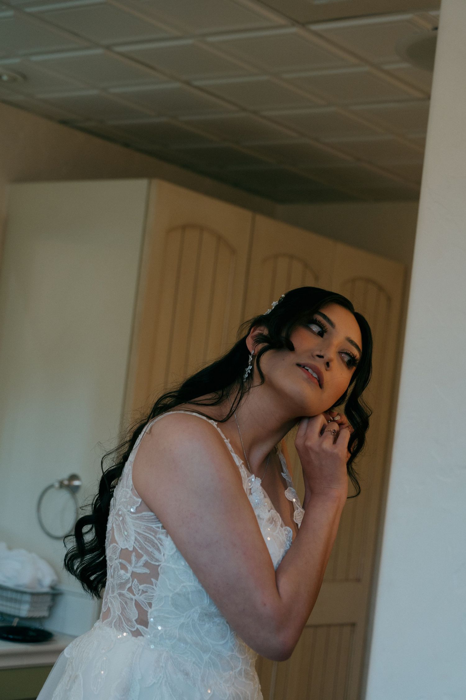 Bride in lace wedding gown adjusting earring while getting ready indoors.