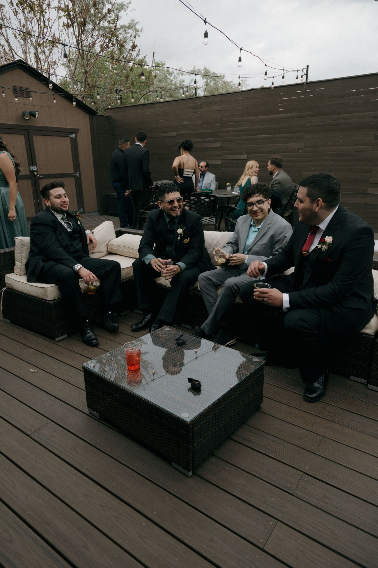 Groomsmen in suits laughing and enjoying drinks together on a wooden deck at a wedding reception.