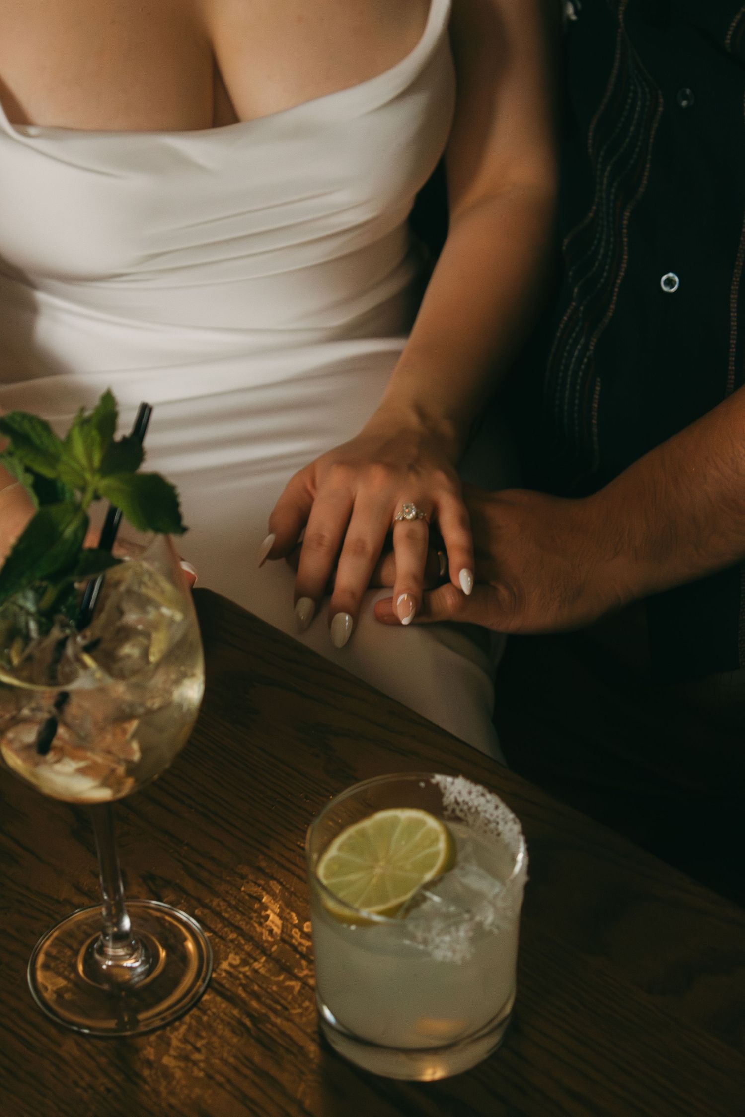 Two cocktails with lime garnish sit on a wooden bar table next to clasped hands.